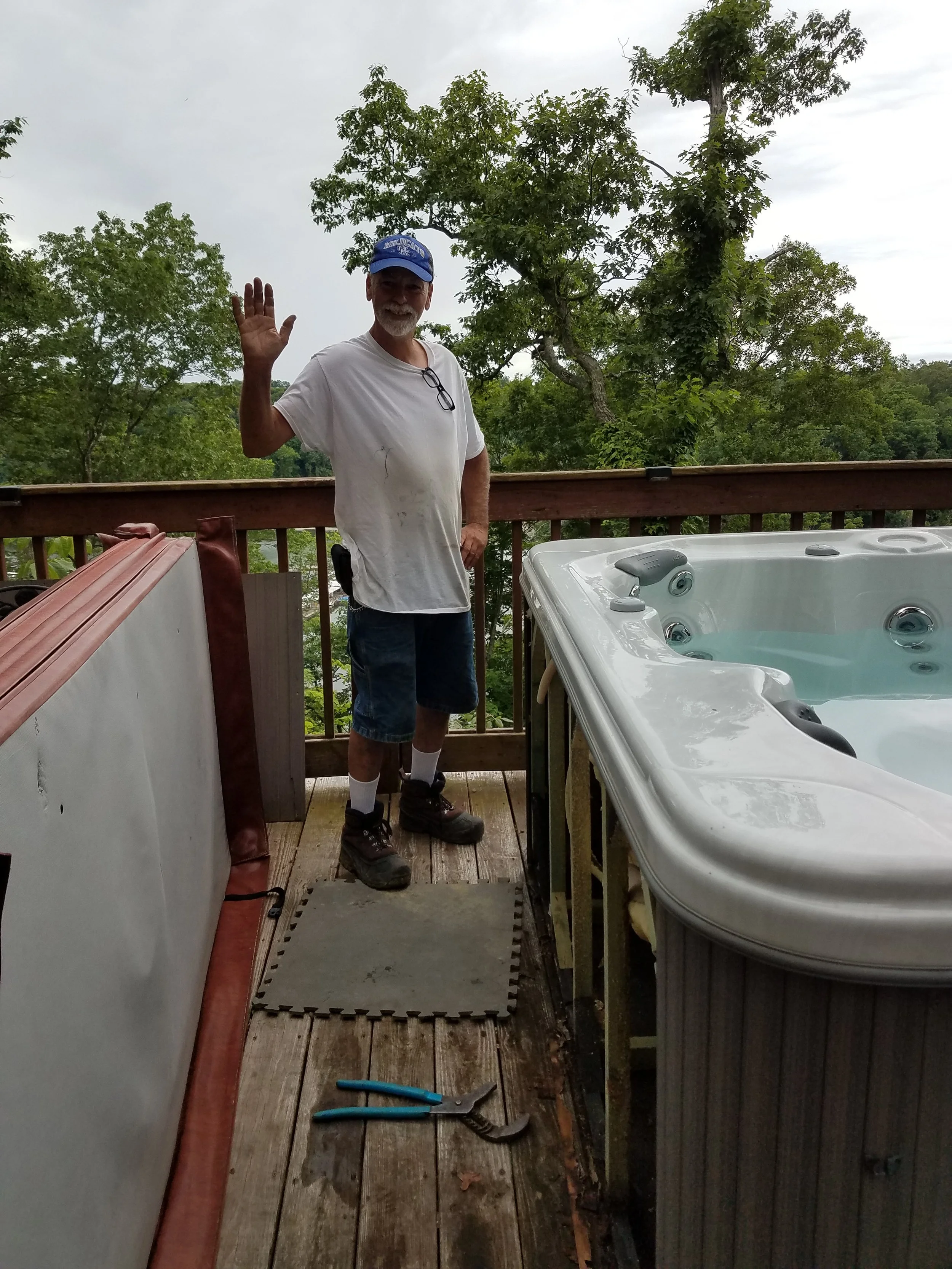 A man standing on a wooden deck, smiling and waving at the camera. He is wearing a white t-shirt, dark shorts, and a blue cap. The deck has various tools and a hot tub, surrounded by trees and an overcast sky.