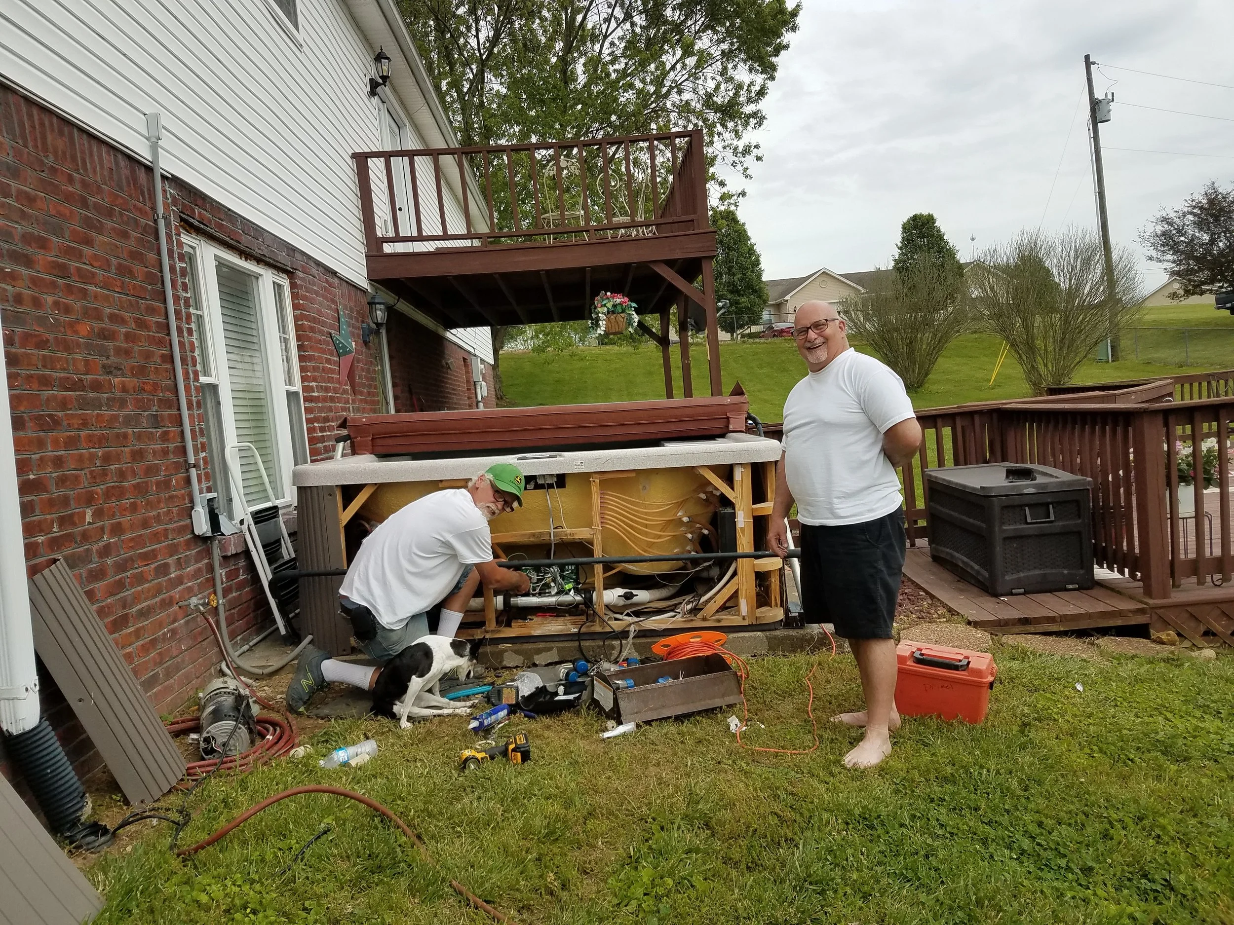 Two men working on a project in a backyard, one bending over and working with tools, and the other standing and smiling. There are plants, a deck, a hot tub, and a house in the background.