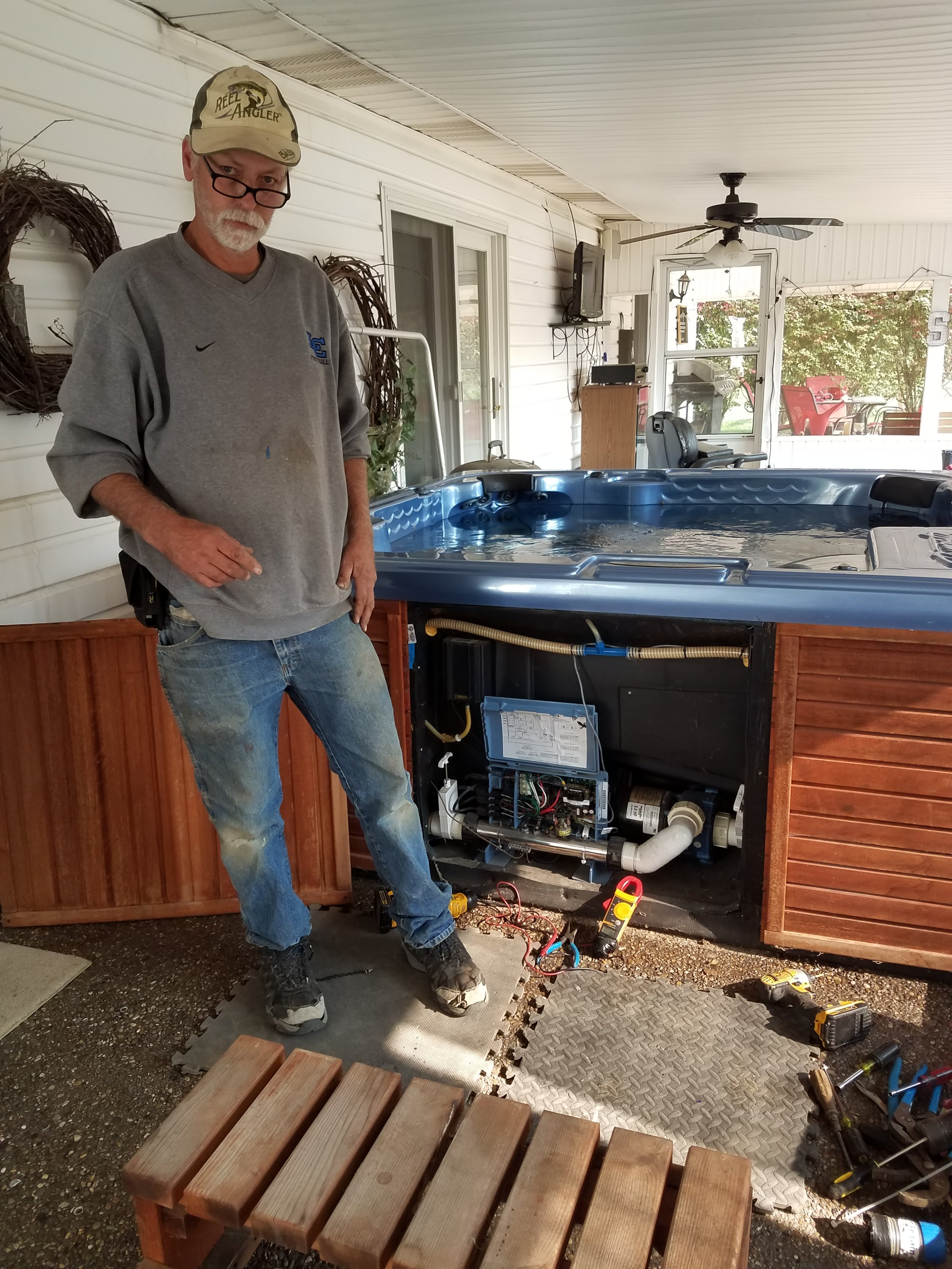 A man with glasses in a gray sweatshirt standing next to a hot tub with the panel removed, revealing electrical and plumbing components, in a porch area with outdoor furniture visible outside.