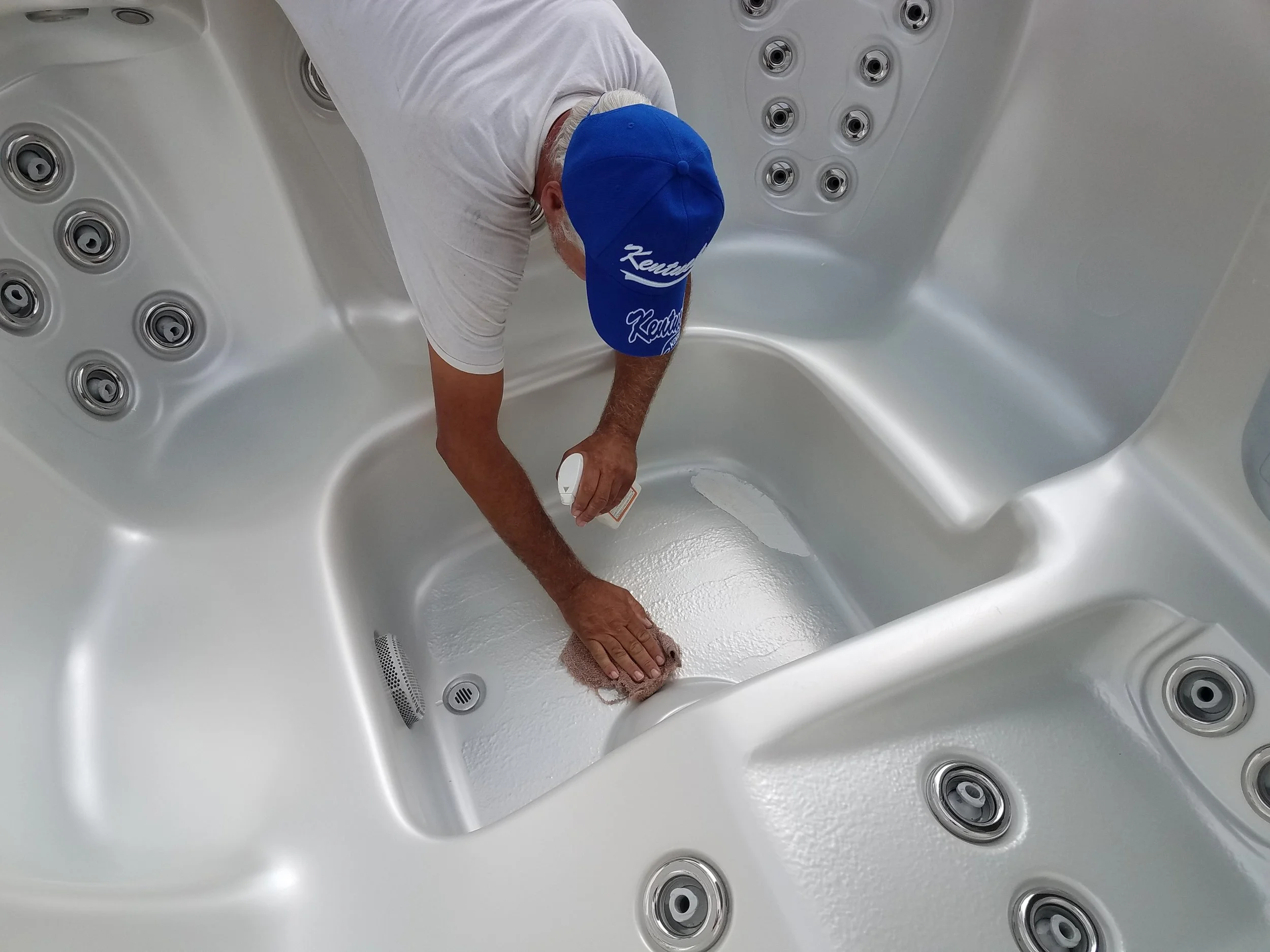 A person with gray hair, wearing a blue cap and white shirt, is cleaning the floor inside a hot tub or jacuzzi with a sponge. The jacuzzi has multiple jets and a smooth white interior.