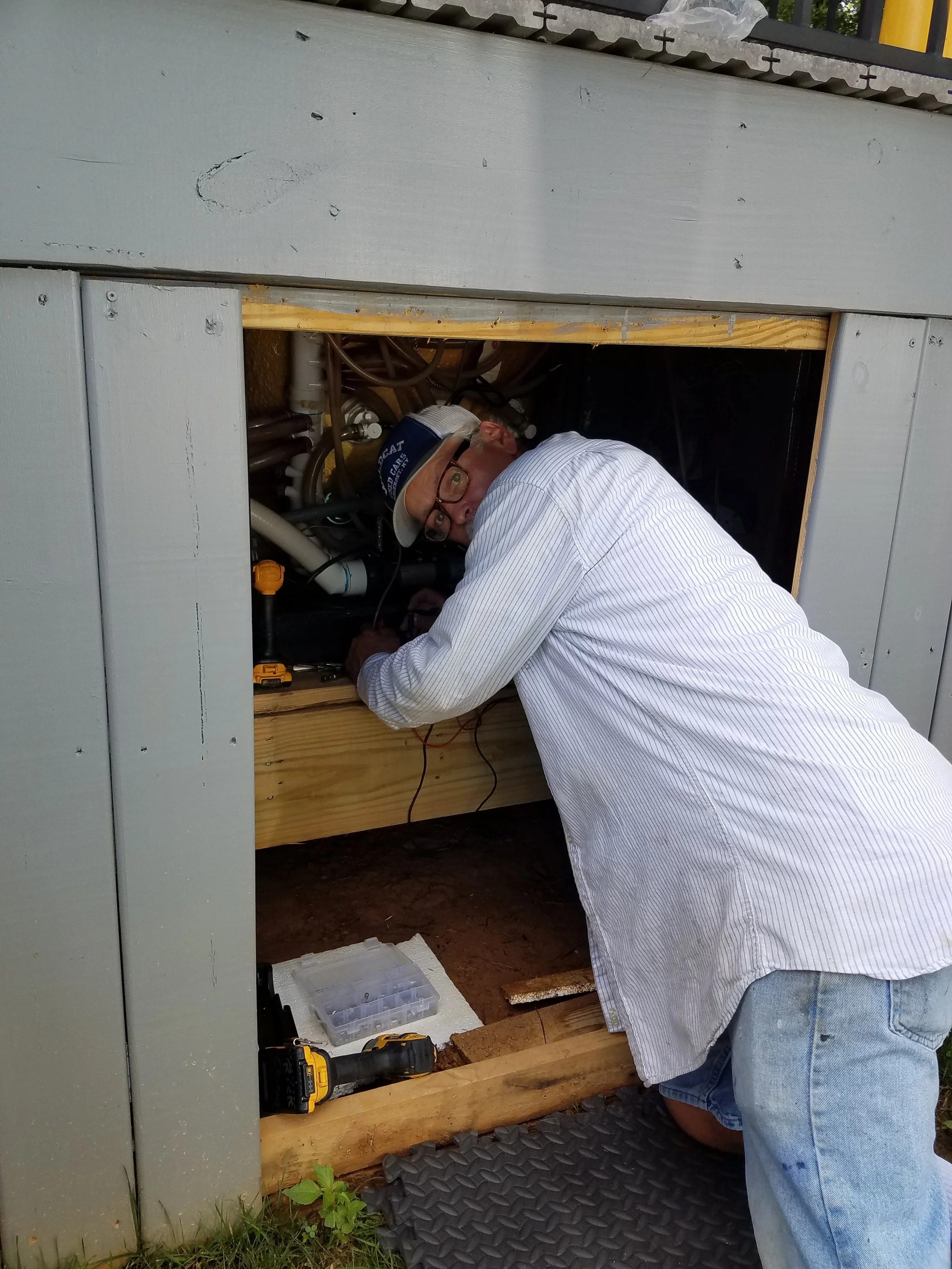 A man with glasses, wearing a cap and a striped shirt, working inside an outdoor electrical or plumbing enclosure with various pipes and wires, using tools.