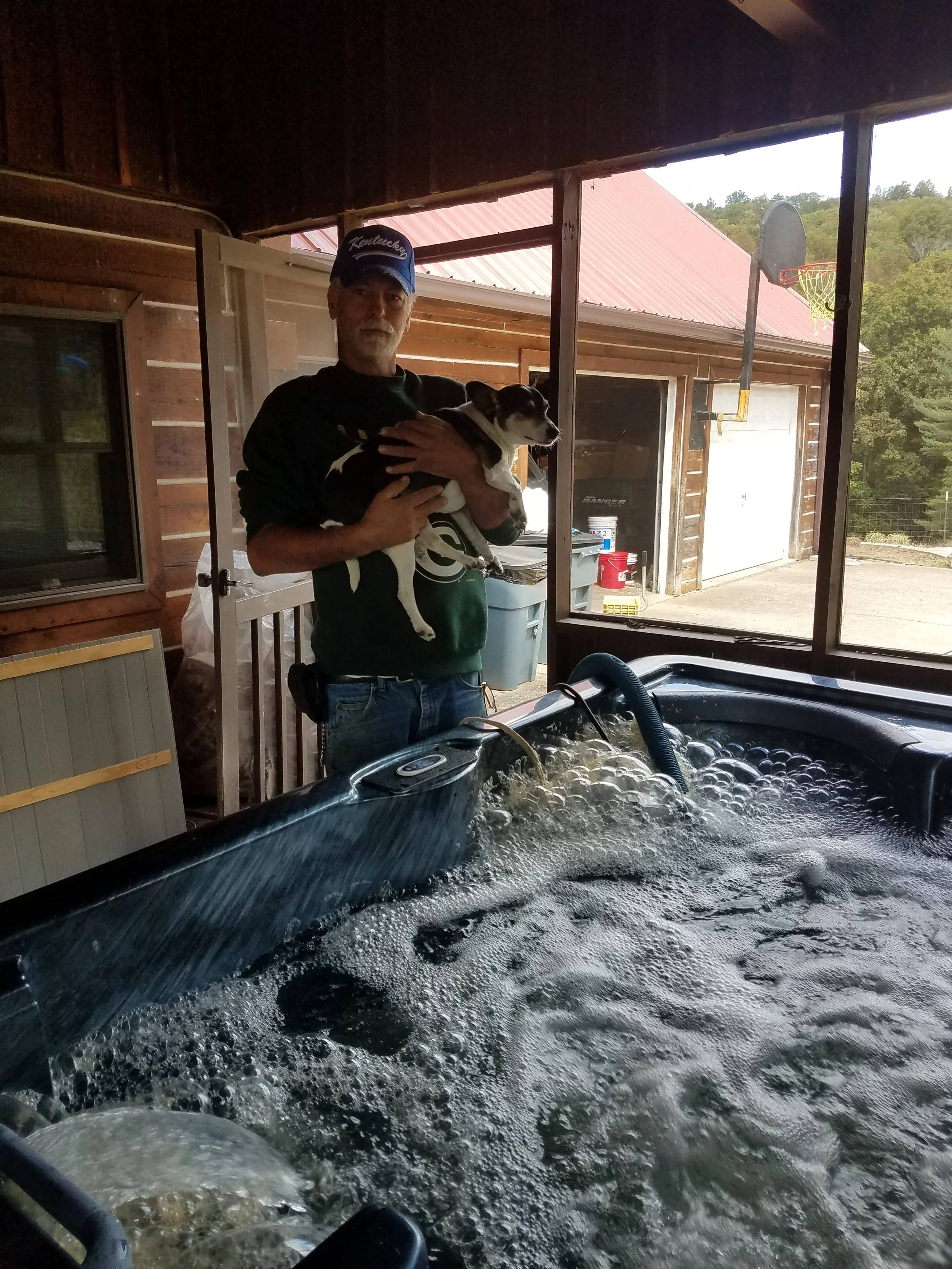 A man with a beard wearing a Kentucky cap holding a small black and white dog inside a wooden structure. The person appears to be next to a hot tub filled with bubbling water, with a backyard visible outside including a basketball hoop and trees.