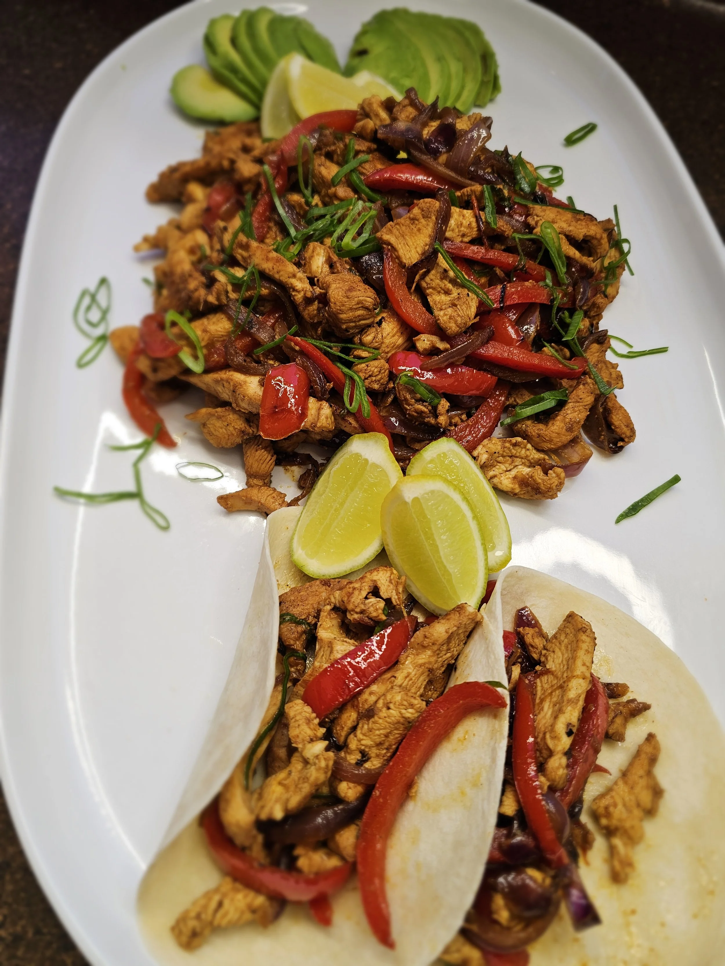 Two chicken fajitas with red bell peppers, onions, and green herbs in flour tortillas, garnished with lemon slices and sliced avocado on a white oval plate.