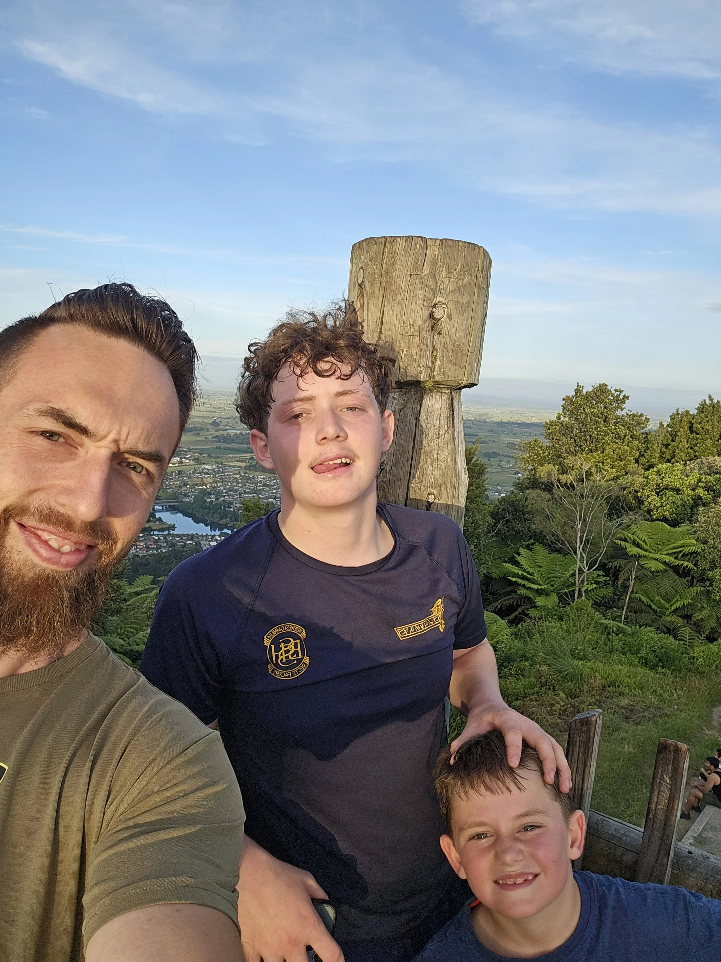 A group of people taking a selfie outdoors on a scenic hilltop with a town, river, and rolling hills in the background. The group includes two adults and three children, all smiling.