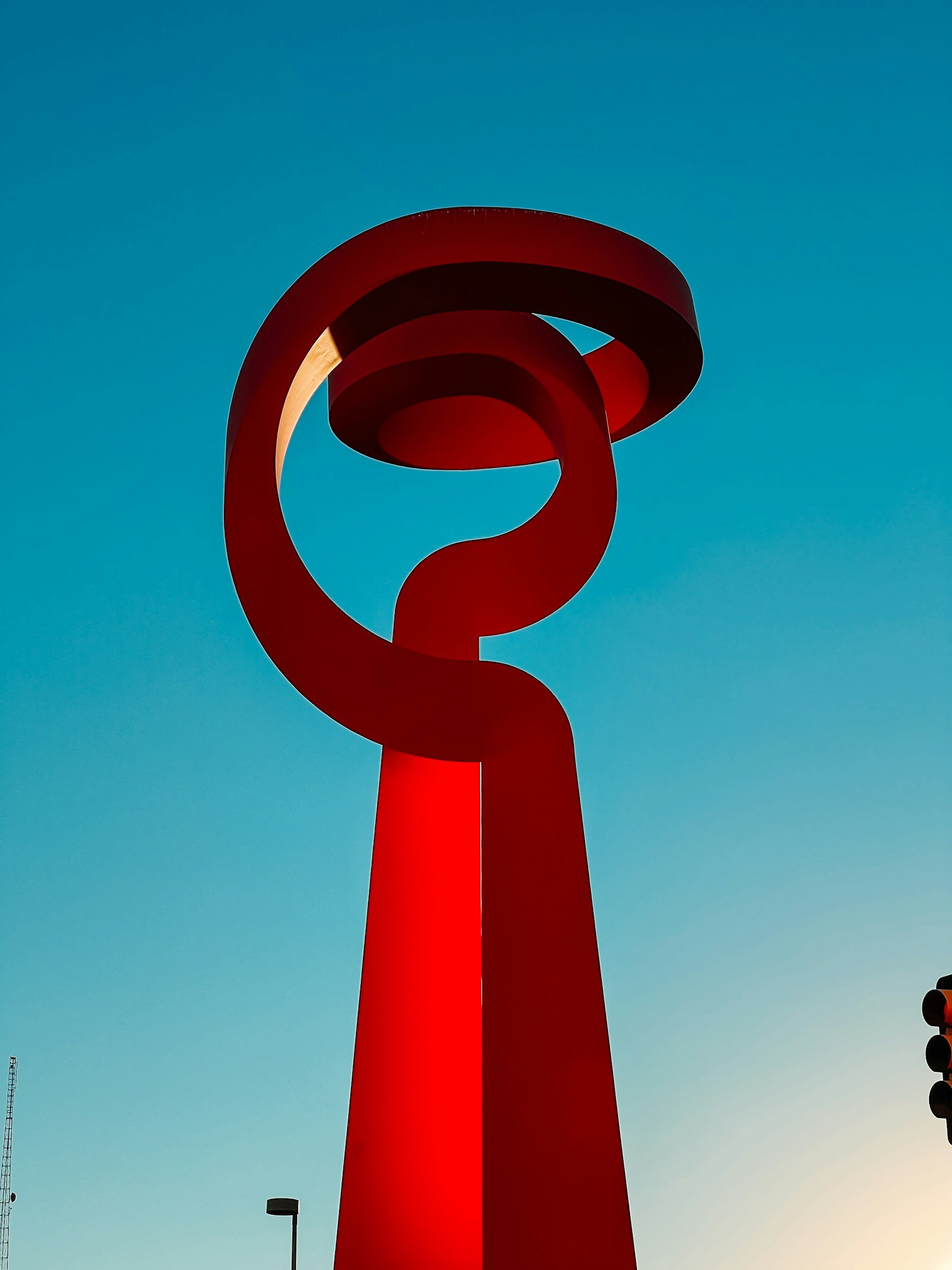 A large red abstract sculpture with multiple loops and bands against a clear blue sky during daytime.