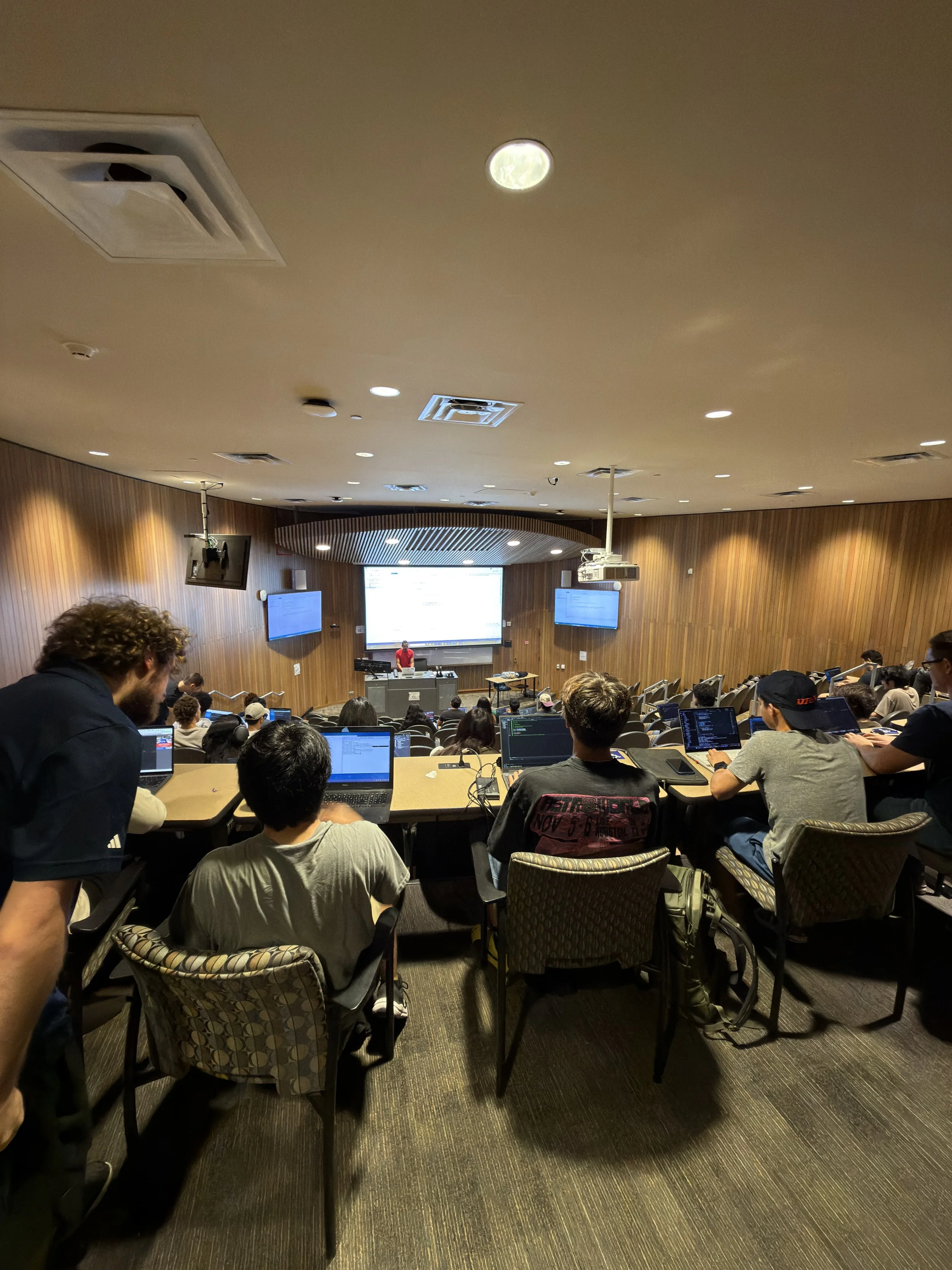 Lecture hall filled with students using laptops, facing a large screen at the front where a presenter in a red shirt is speaking.