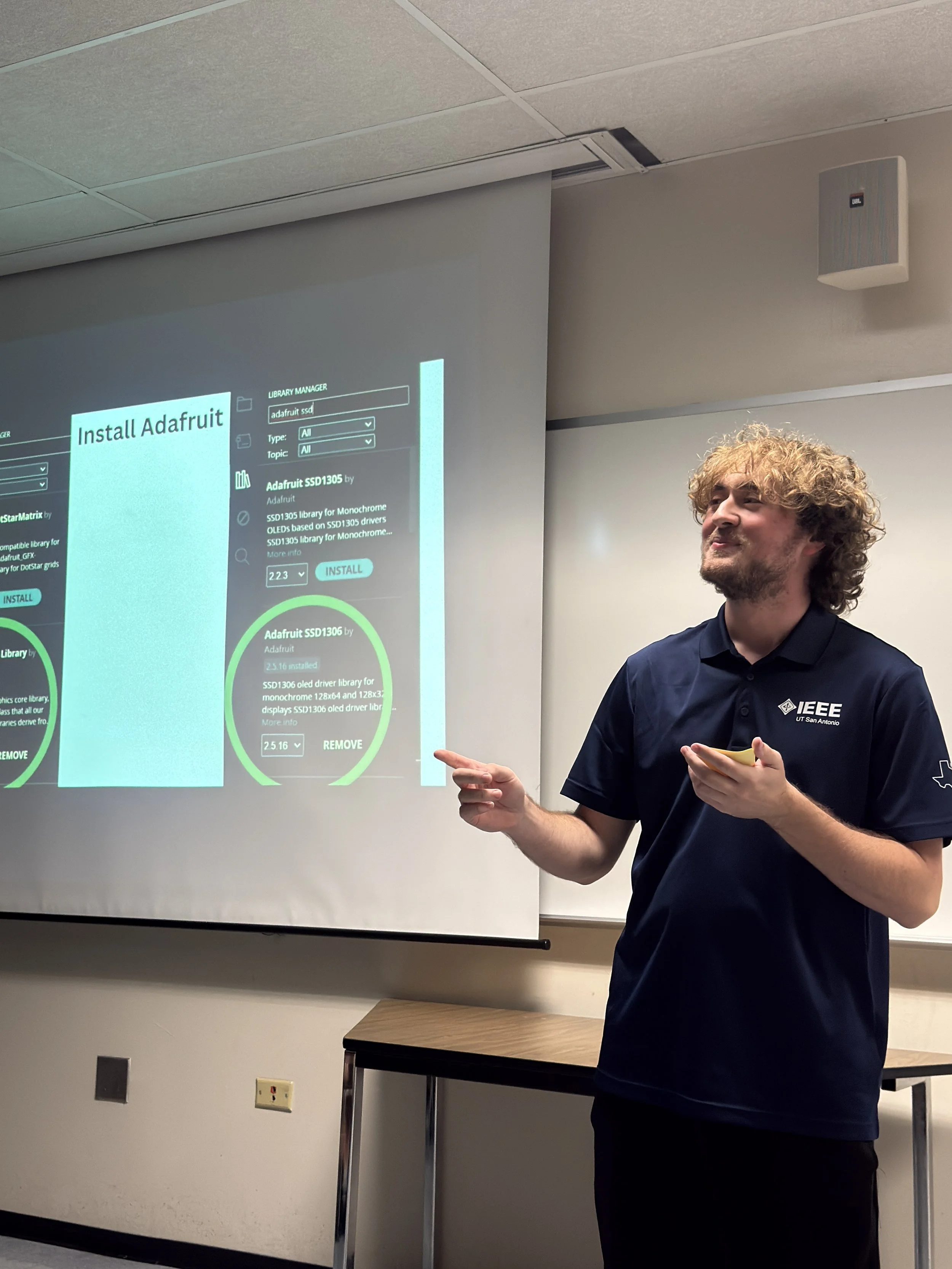 A man with curly blonde hair and a beard wearing a navy blue polo shirt with an IEEE logo is giving a presentation in a classroom. He is pointing at a projection screen showcasing a software interface related to installing Adafruit hardware.