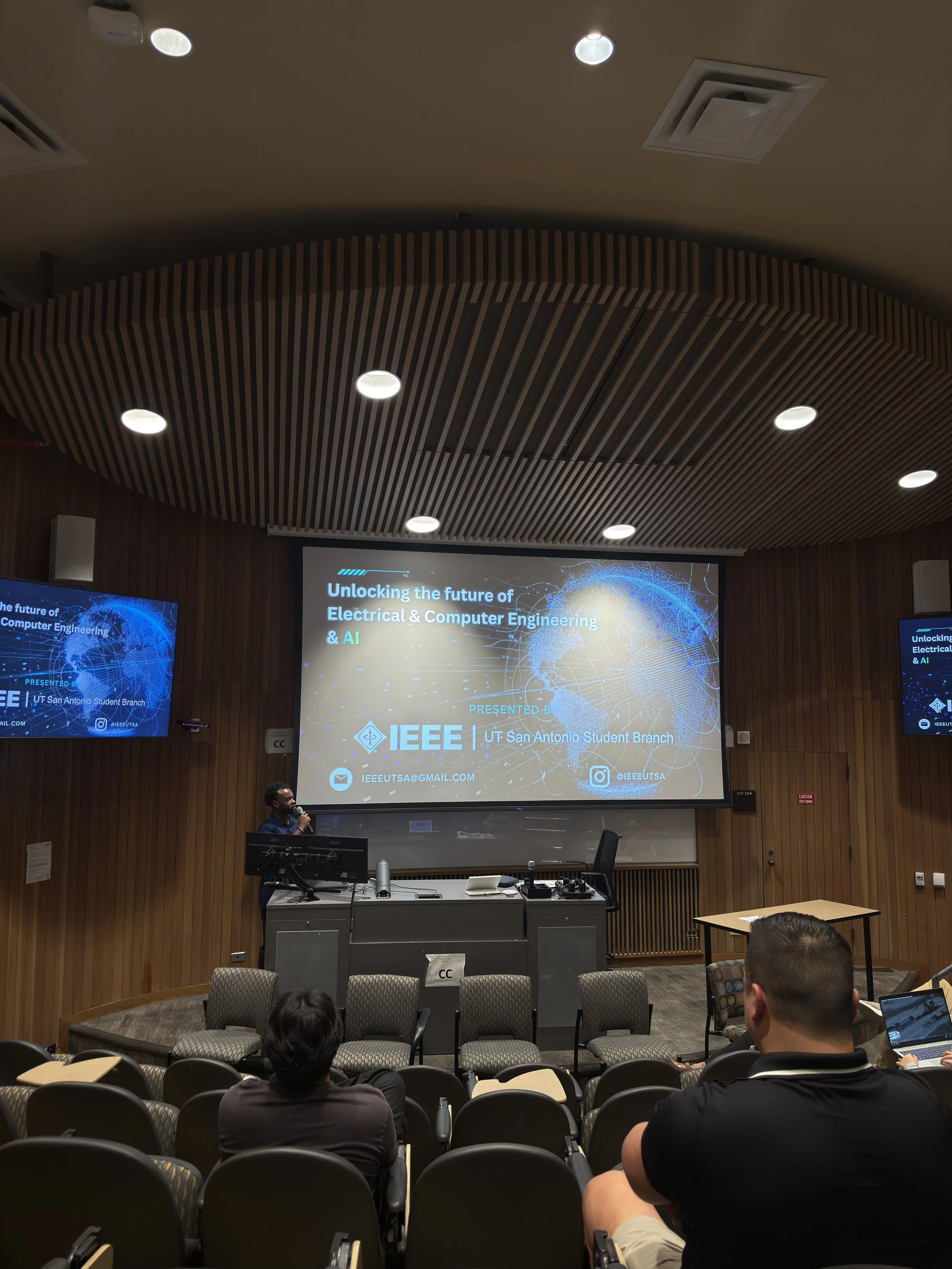 A lecture hall or conference room with a presentation on a screen about the future of electrical and computer engineering and AI by IEEE UT San Antonio Student Branch. There are several attendees seated, including a woman at the podium and a man with a microphone, with a wooden wall and ceiling design.