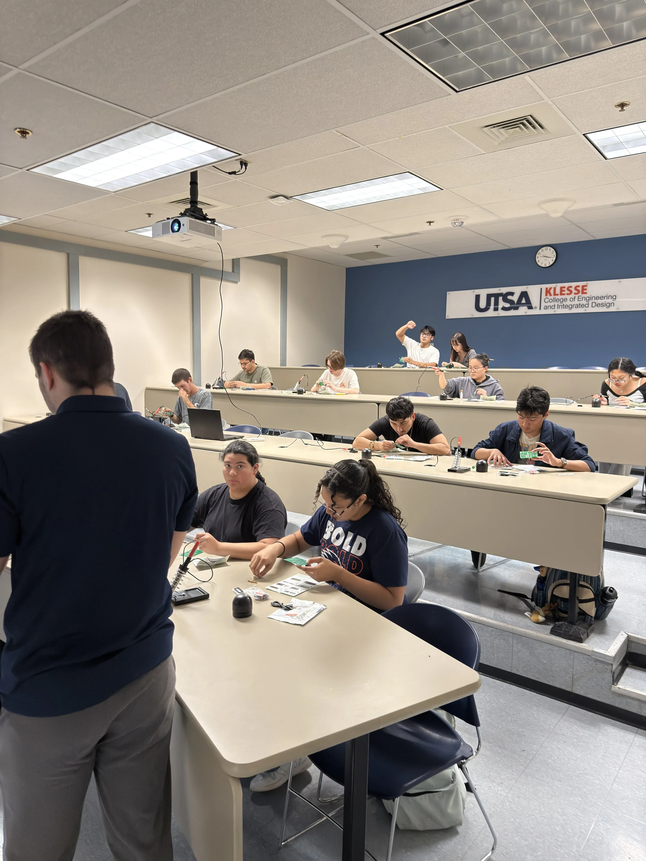 Students working on electronics projects in a classroom at UTSA Klesse College of Engineering. Some students are seated at desks with electronic components, while others are standing or supervising.