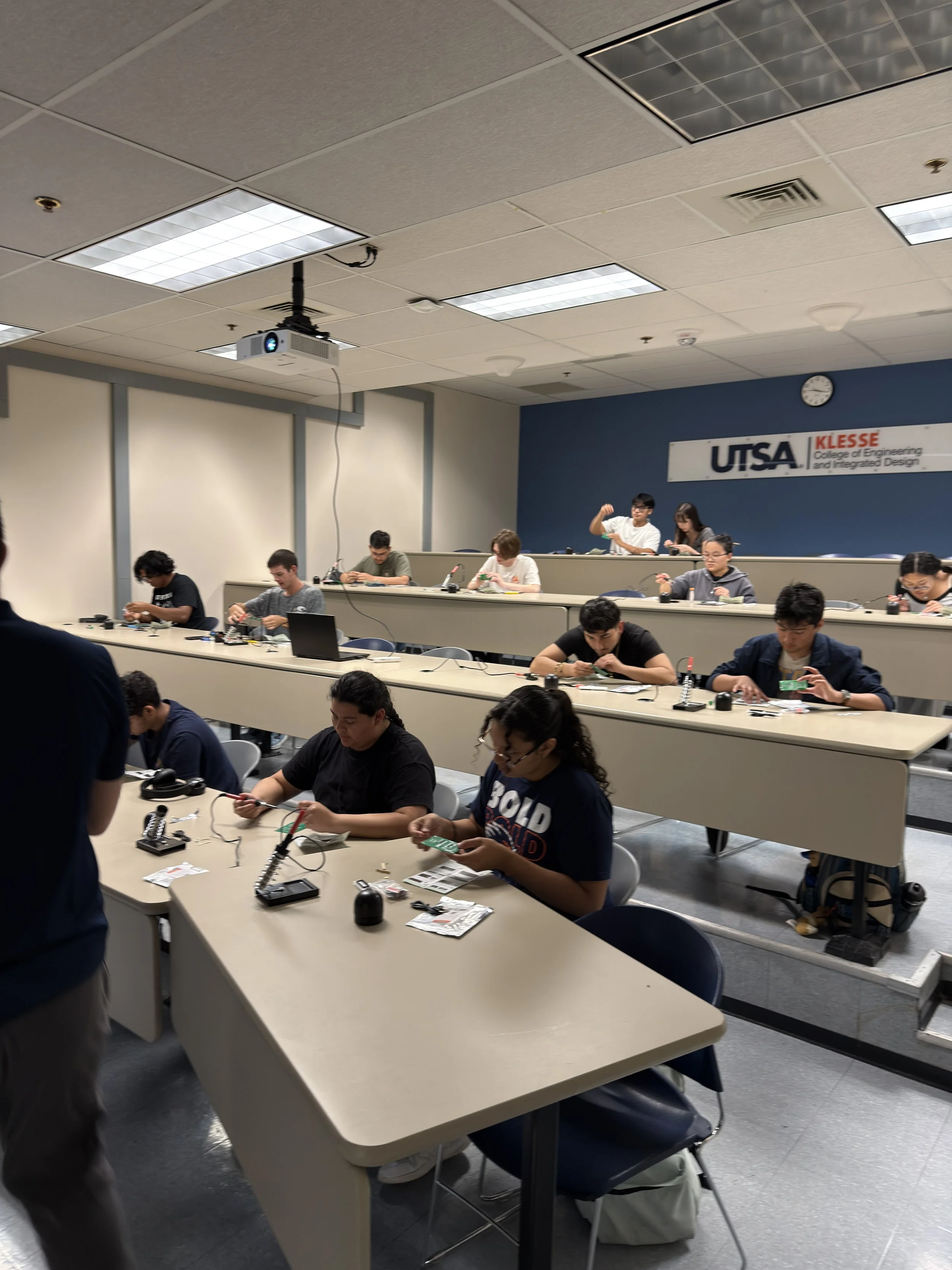 A classroom with multiple students working on electronics projects at desks, with a sign on the wall reading 'UTSA KLESSE College of Engineering and Integrated Design' and a clock showing 2:25.