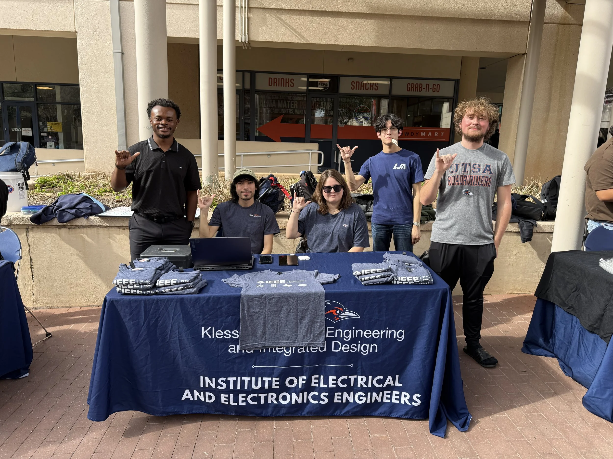 Group of five students at a table outside the Institute of Electrical and Electronics Engineers, showing promotional shirts and backpacks.