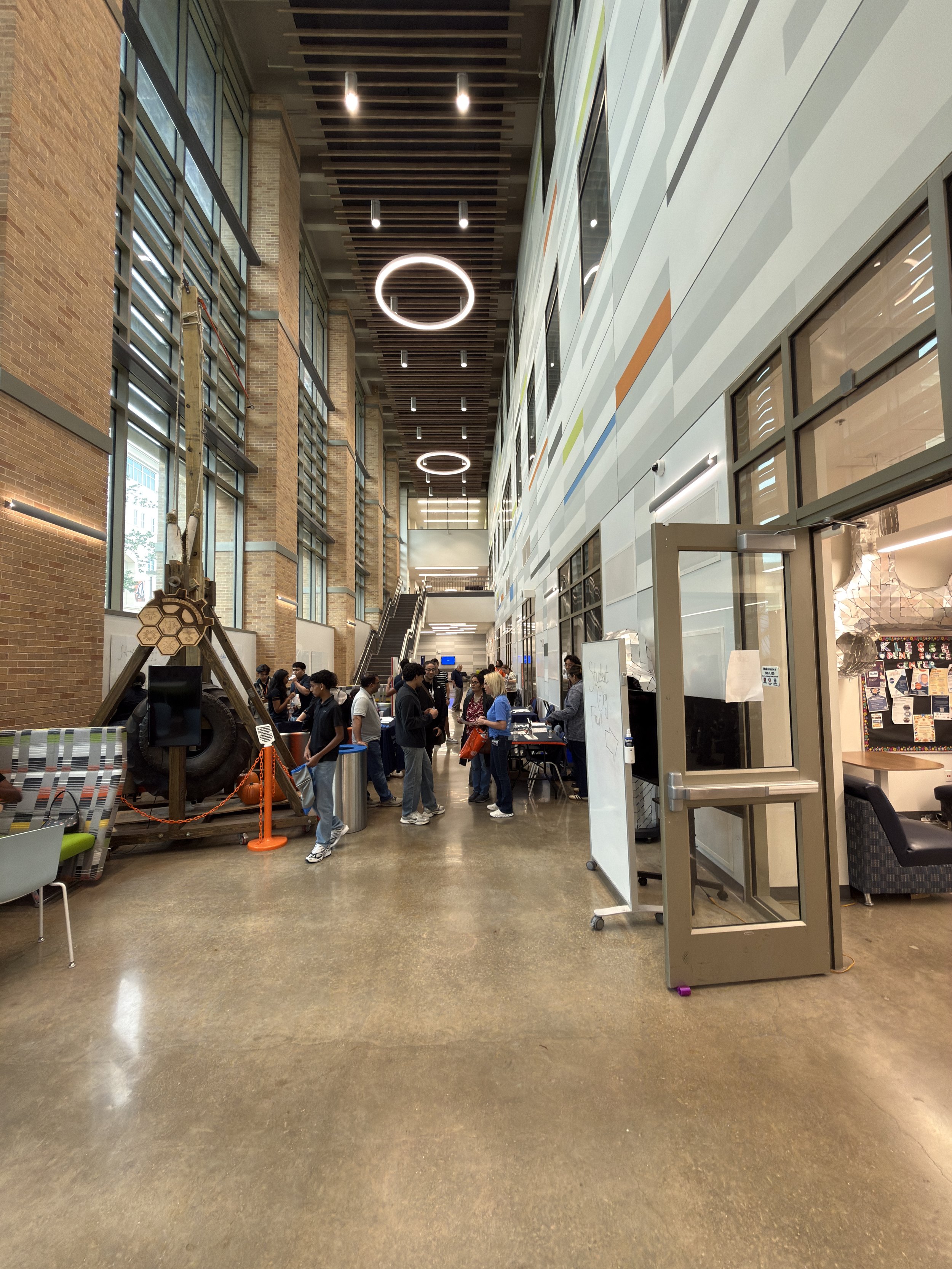 Scene inside a modern building with high ceilings, large windows, and a group of people gathered around tables and standing in line near a registration or check-in desk. There is a display with a tire and a honeycomb structure, and an elevator door to the right.
