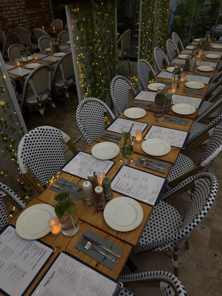 A long dining table set for a restaurant or event with plates, menus, water glasses with mint leaves, candles, and condiment containers, decorated with fairy lights and surrounded by checkered chairs.