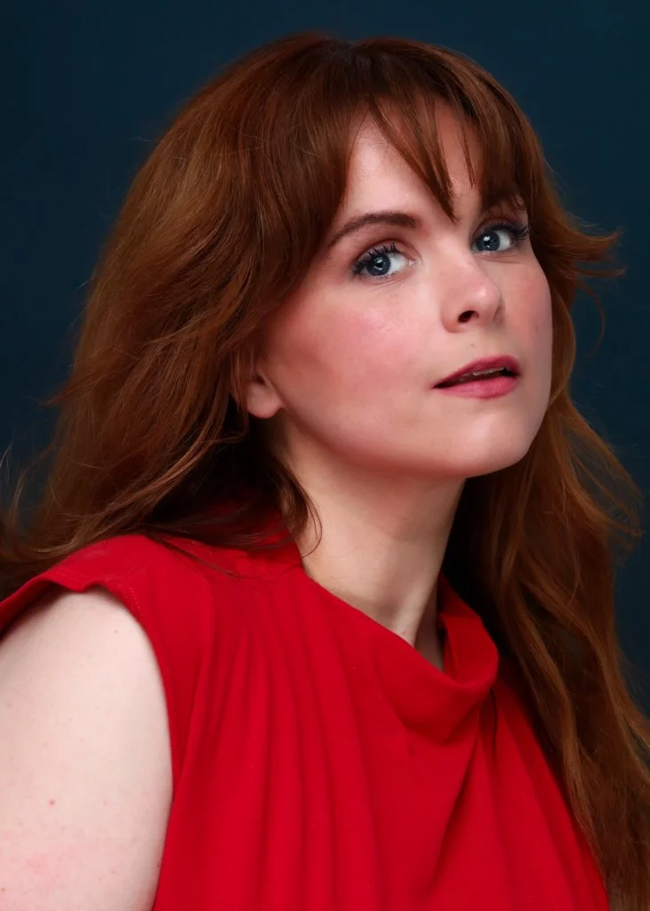 Blaire Erskine, comedy writer and actor, in a red top looking over her shoulder against a dark blue background.