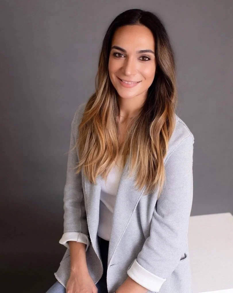 A woman with long, wavy brown hair and light skin, smiling at the camera, wearing a light gray blazer and white top, seated against a plain gray background.