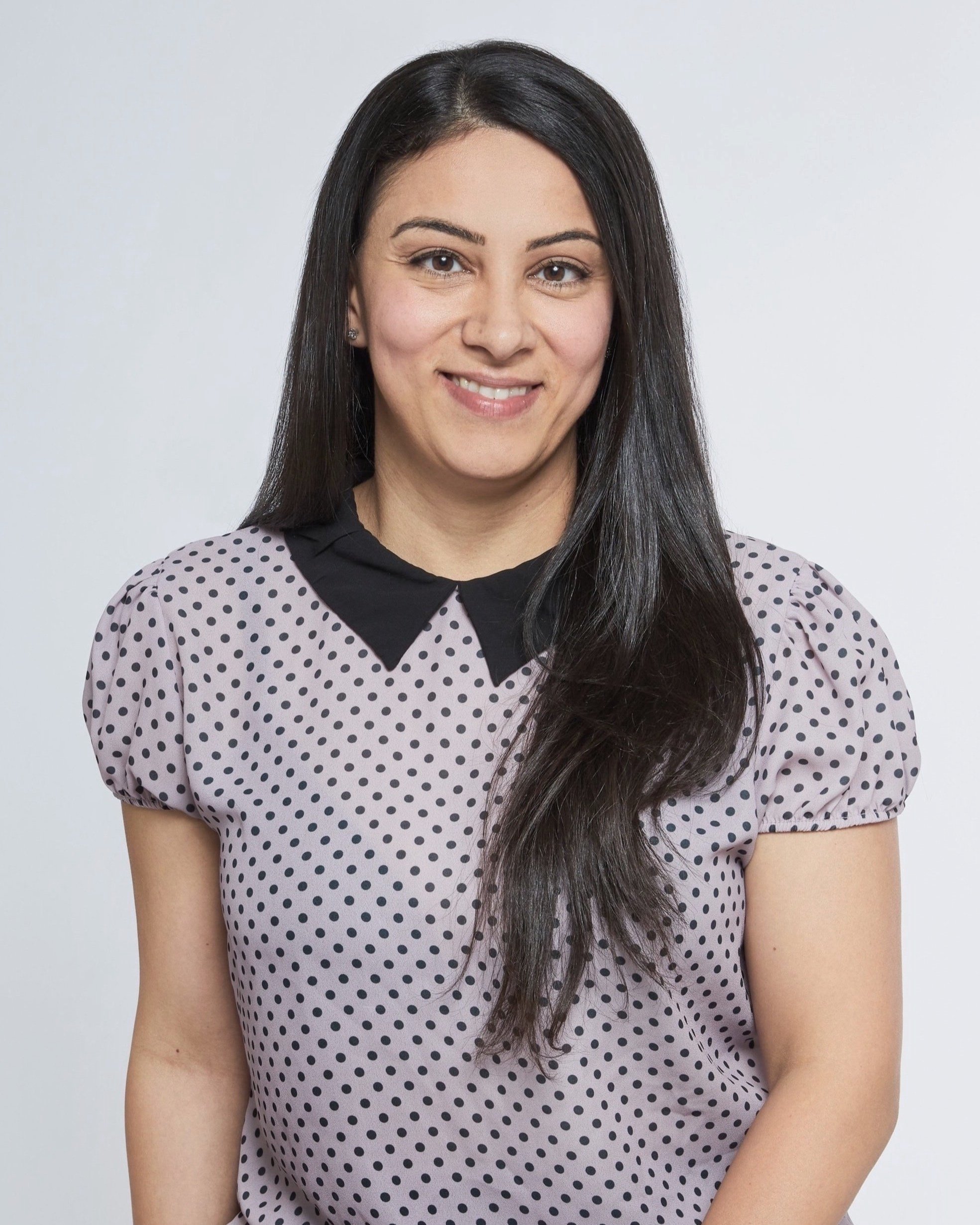 A woman with long dark hair wearing a lilac blouse with black polka dots and a black collar, smiling against a plain white background.