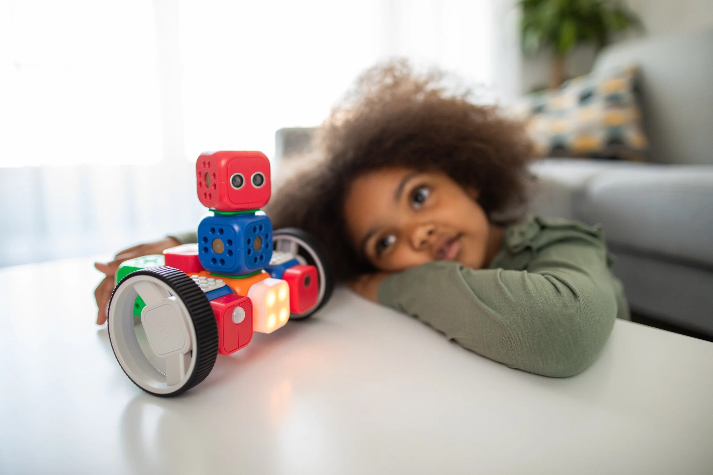 A young girl with curly hair reclining on a table, resting her head on her arm, and looking at a colorful robot toy with wheels and lights.