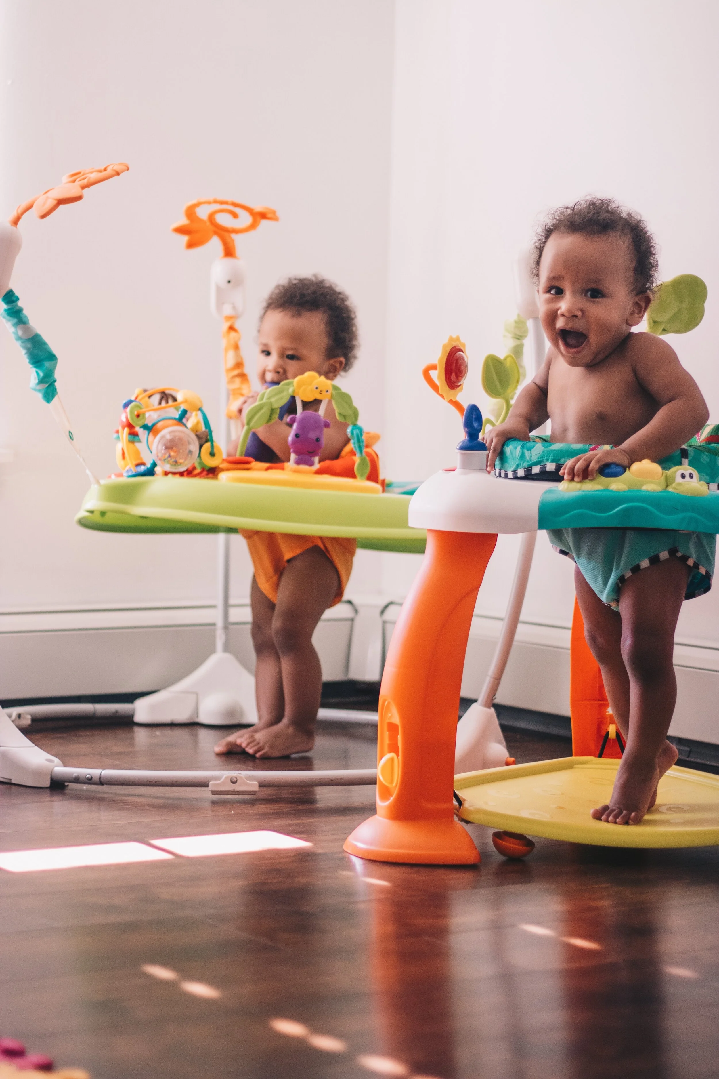 Two young children playing in baby walkers in a room with hardwood floors and white walls.