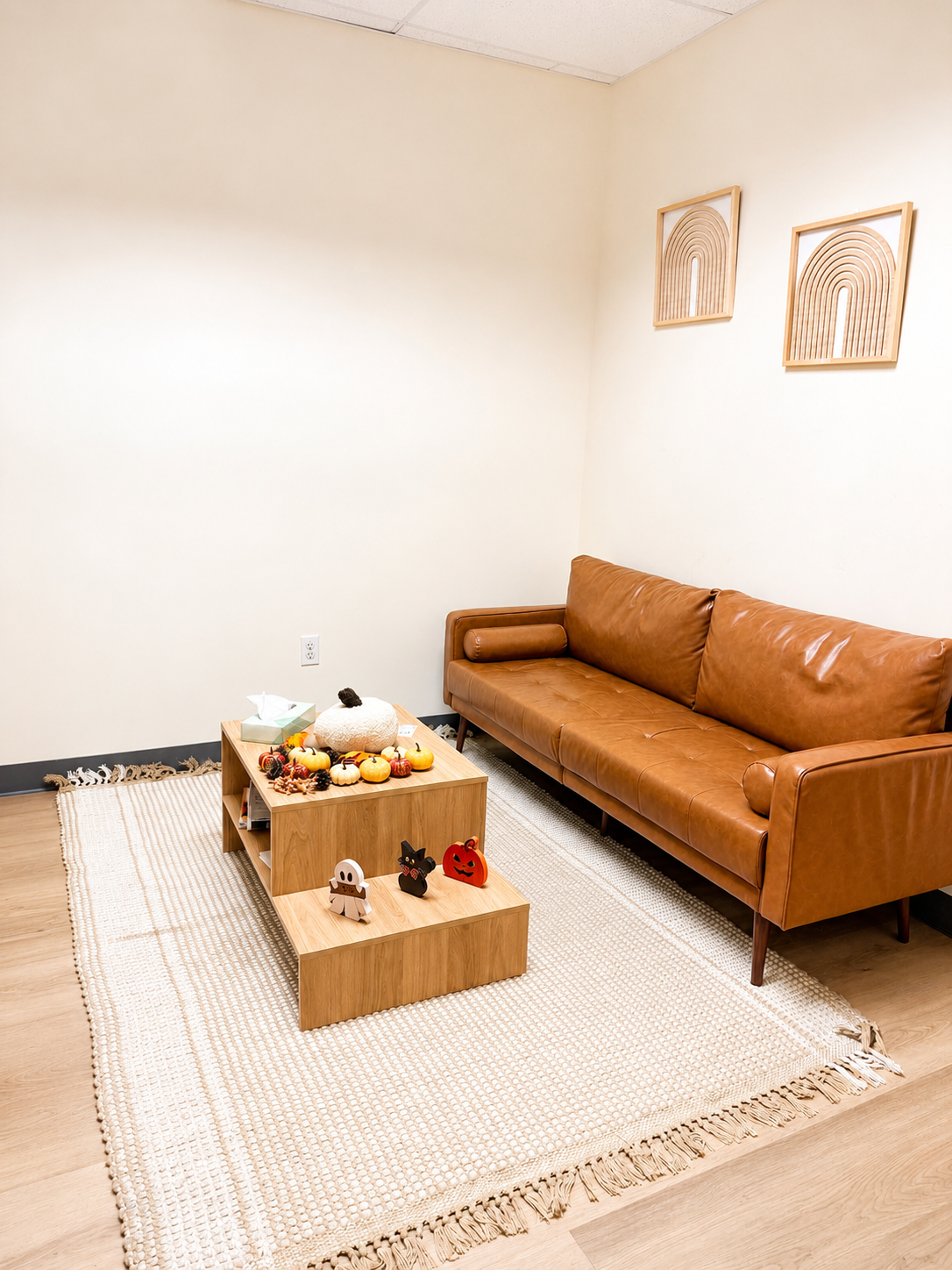 A cozy room with a tan leather sofa, a wooden coffee table decorated with autumn Halloween themed decorations, and two framed abstract rainbow artworks on the wall. The room has a cream-colored area rug and a light wood floor.