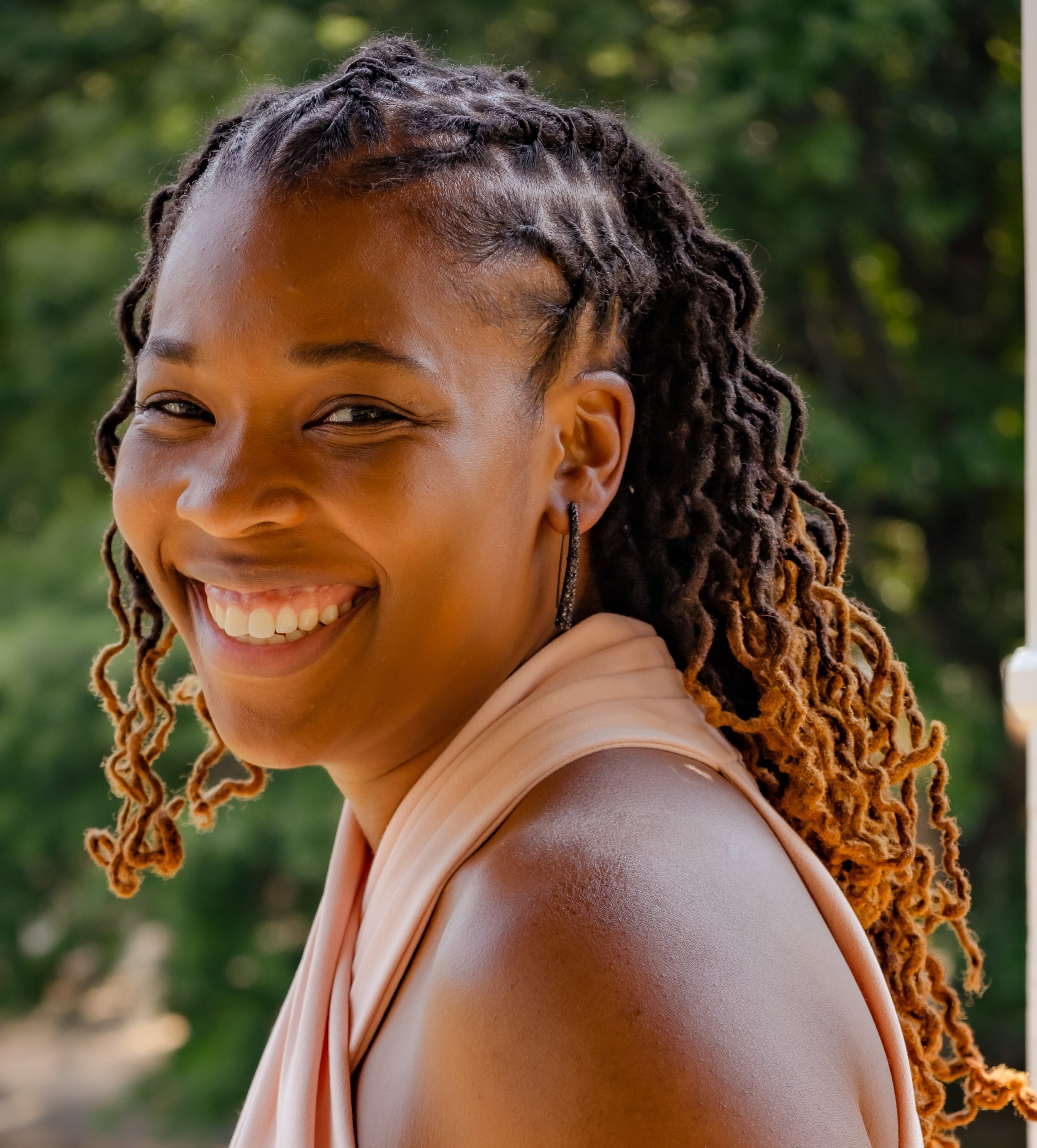 Close-up of a smiling woman with dreadlocks, wearing a light pink top with one shoulder exposed. The background features a blurred green outdoor setting.