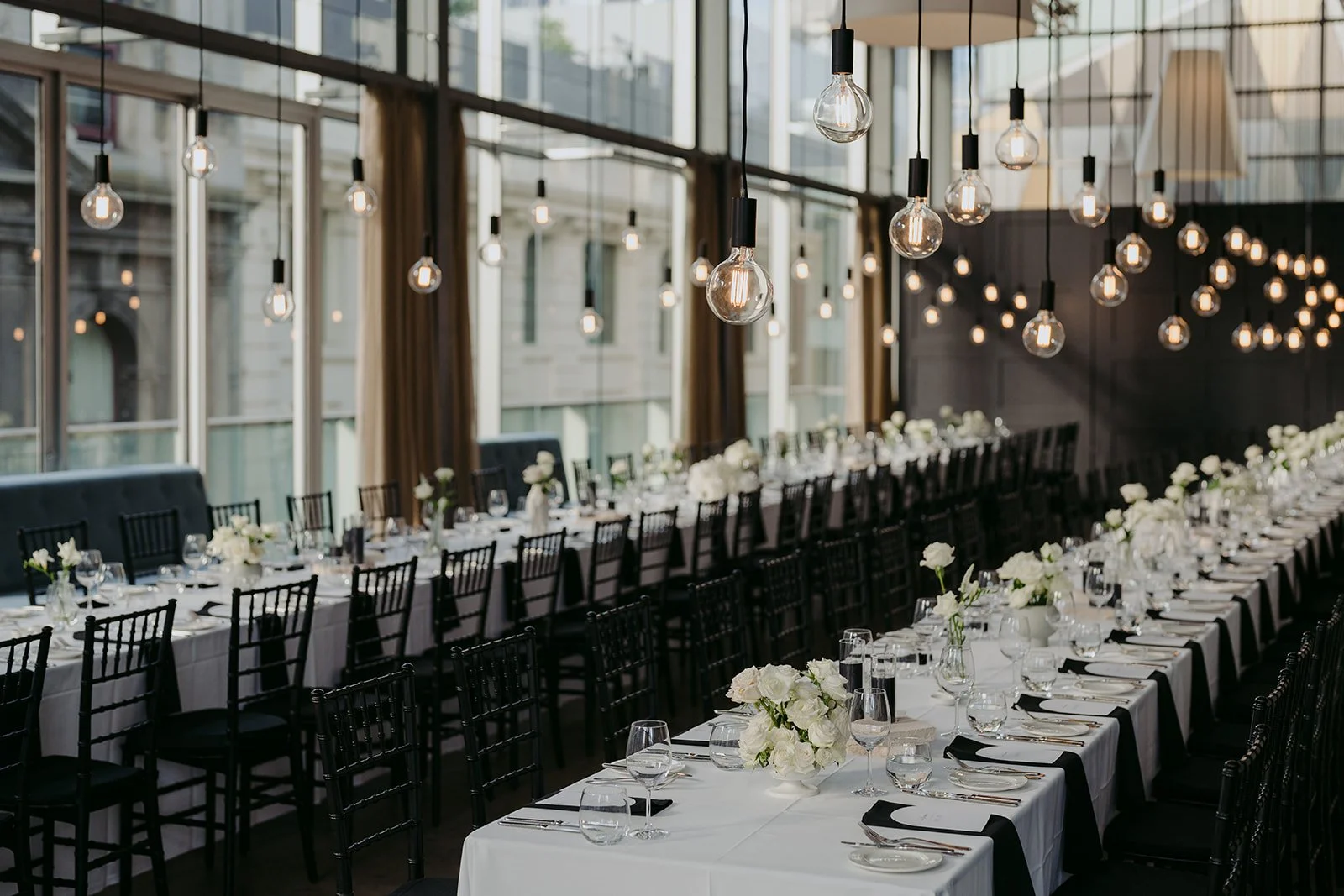 An elegant banquet hall with long tables set for a formal event, decorated with white flowers, glassware, and black chairs. The room has large windows, modern hanging light bulbs, and a high glass ceiling, creating a bright, open atmosphere.