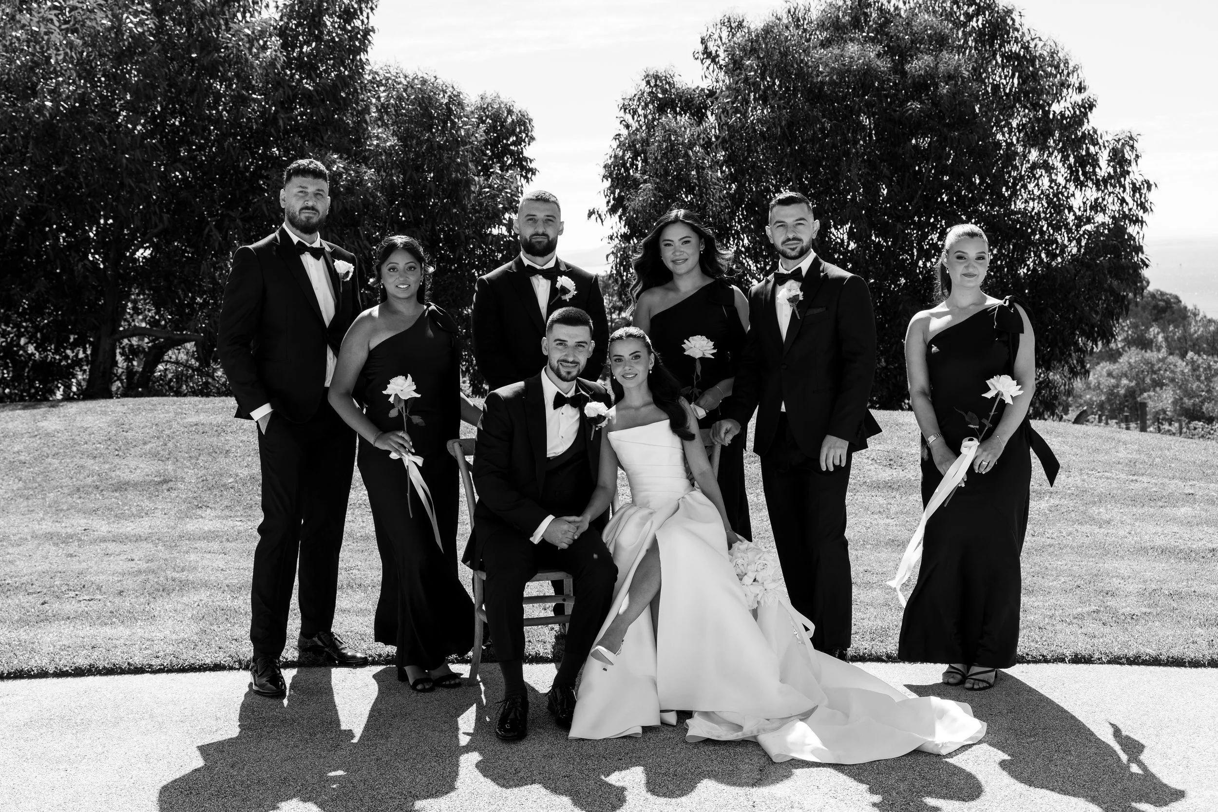 A group of ten people, including women in elegant black dresses and men in tuxedos, posing outdoors in a grassy area with large trees in the background. Two women are seated, one in a white dress and the other in black, with the remaining standing around them. The group appears to be at a formal event or wedding.