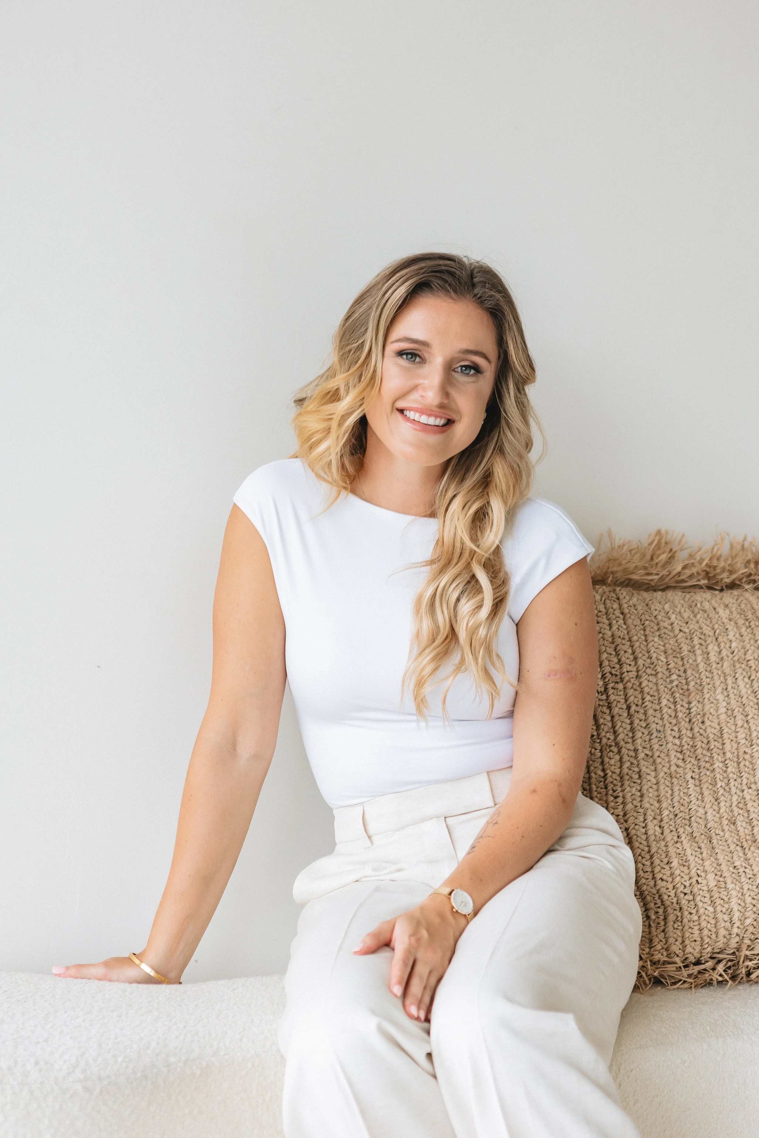 A young woman with long wavy blonde hair, wearing a white top and cream-colored pants, sitting on a beige textured surface with a cozy blanket behind her, smiling at the camera against a plain off-white wall.