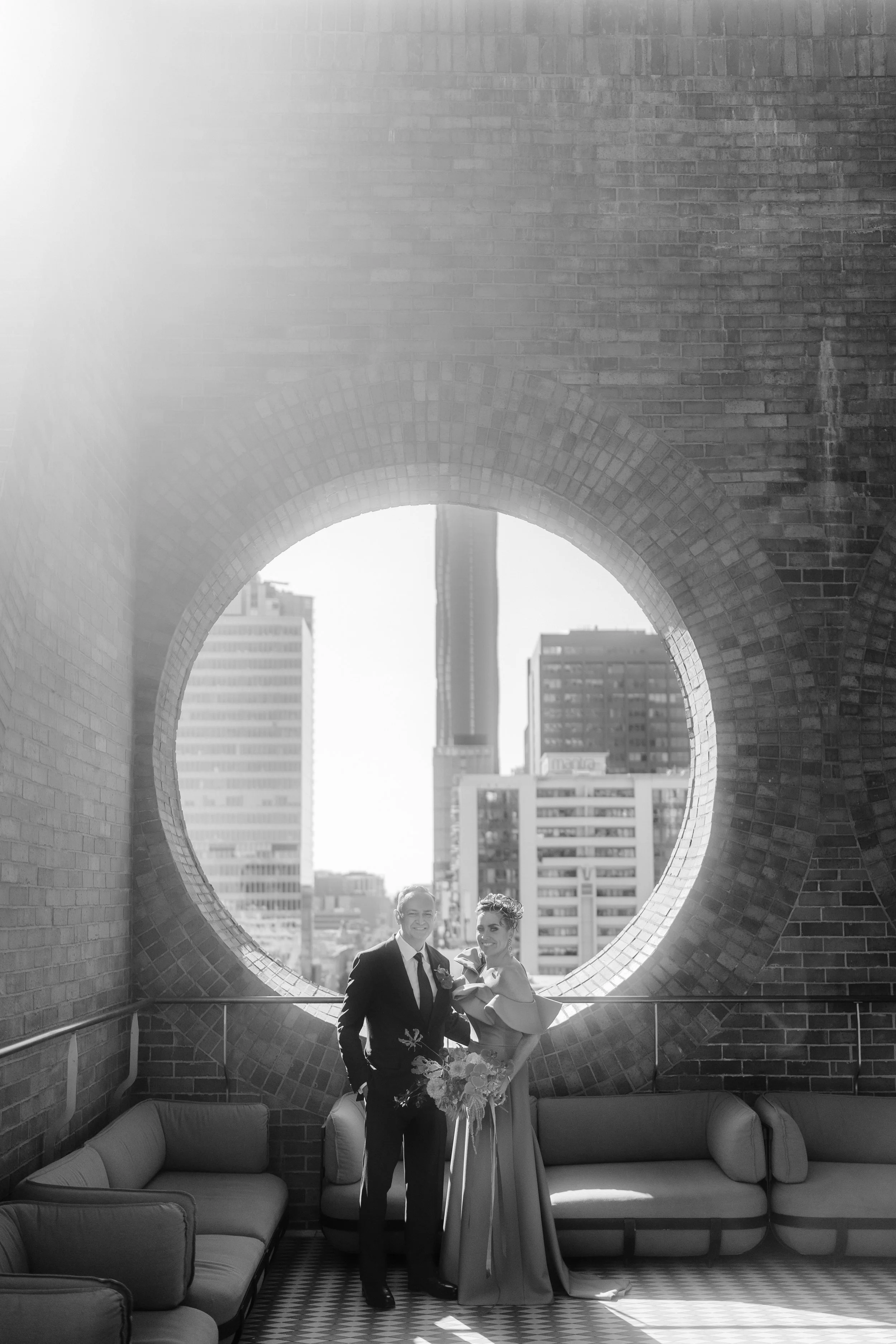 A bride and groom standing together inside a circular brick structure with large windows showing city buildings in the background. The couple is smiling, and the bride holds a bouquet. There are couches around them on a patterned floor.