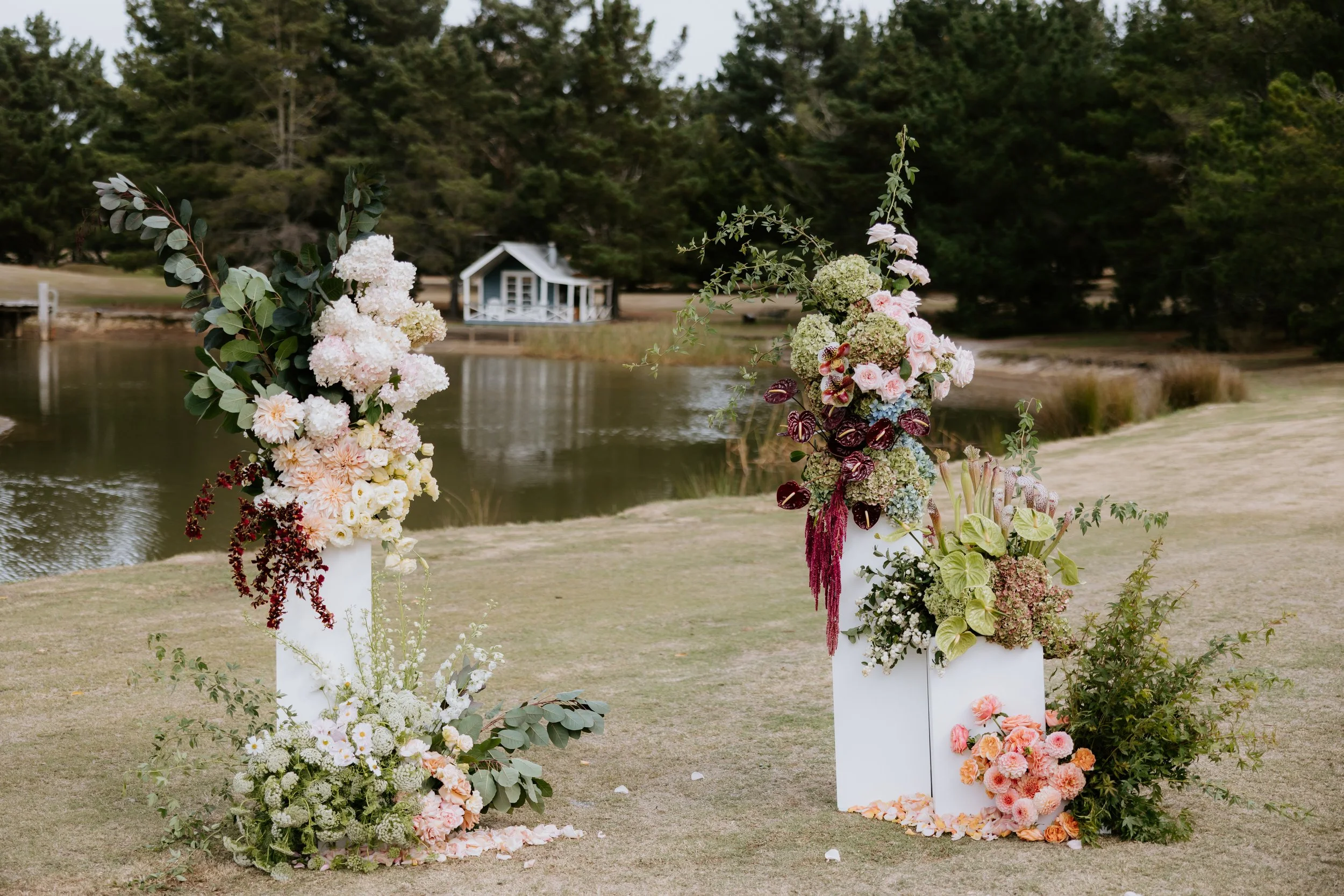 Colorful floral arrangements on white pedestals are set up outdoors near a water body with trees and a small building in the background.