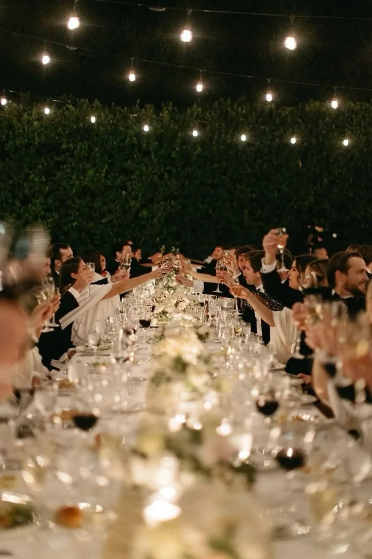 People sitting at a long dinner table outdoors at night, raising glasses for a toast, decorated with flowers, under hanging string lights.