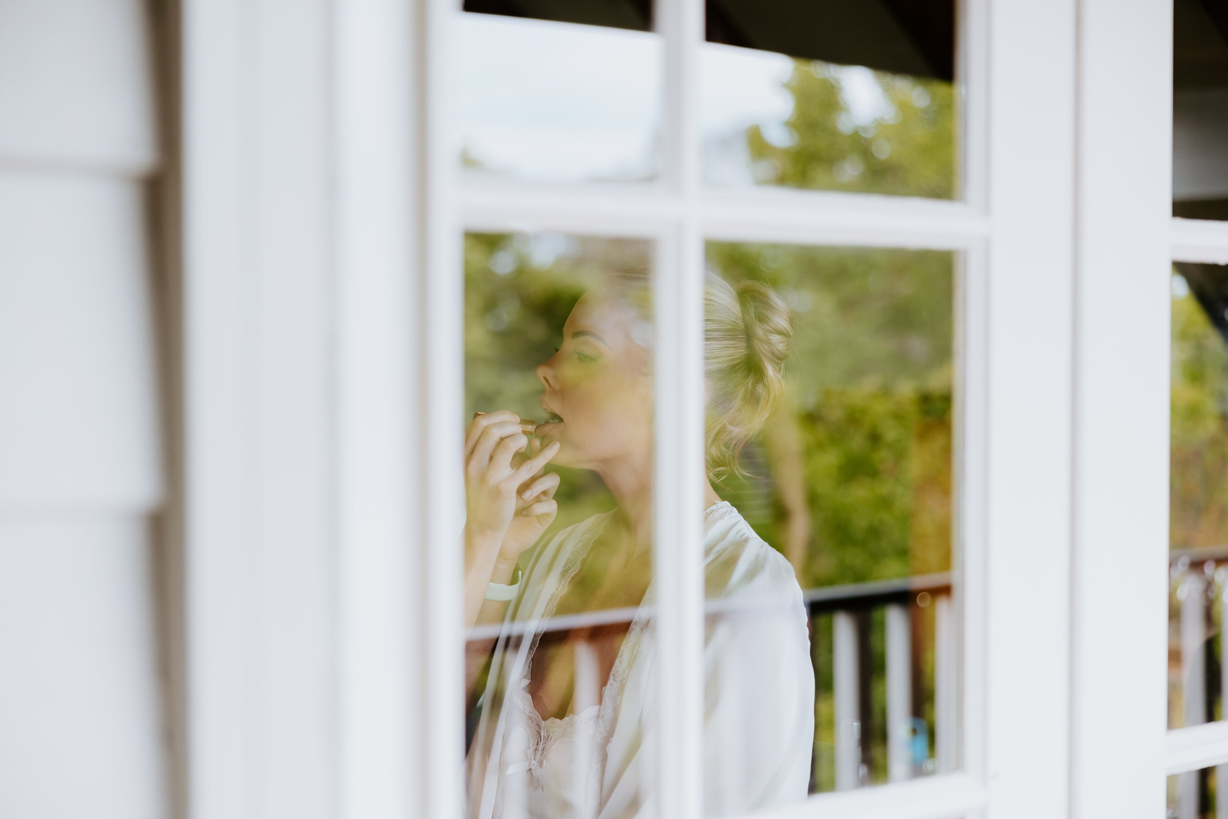 A woman with blonde hair styled in an updo, wearing a white dress, seen through a window with white frames, outdoors with greenery in the background.
