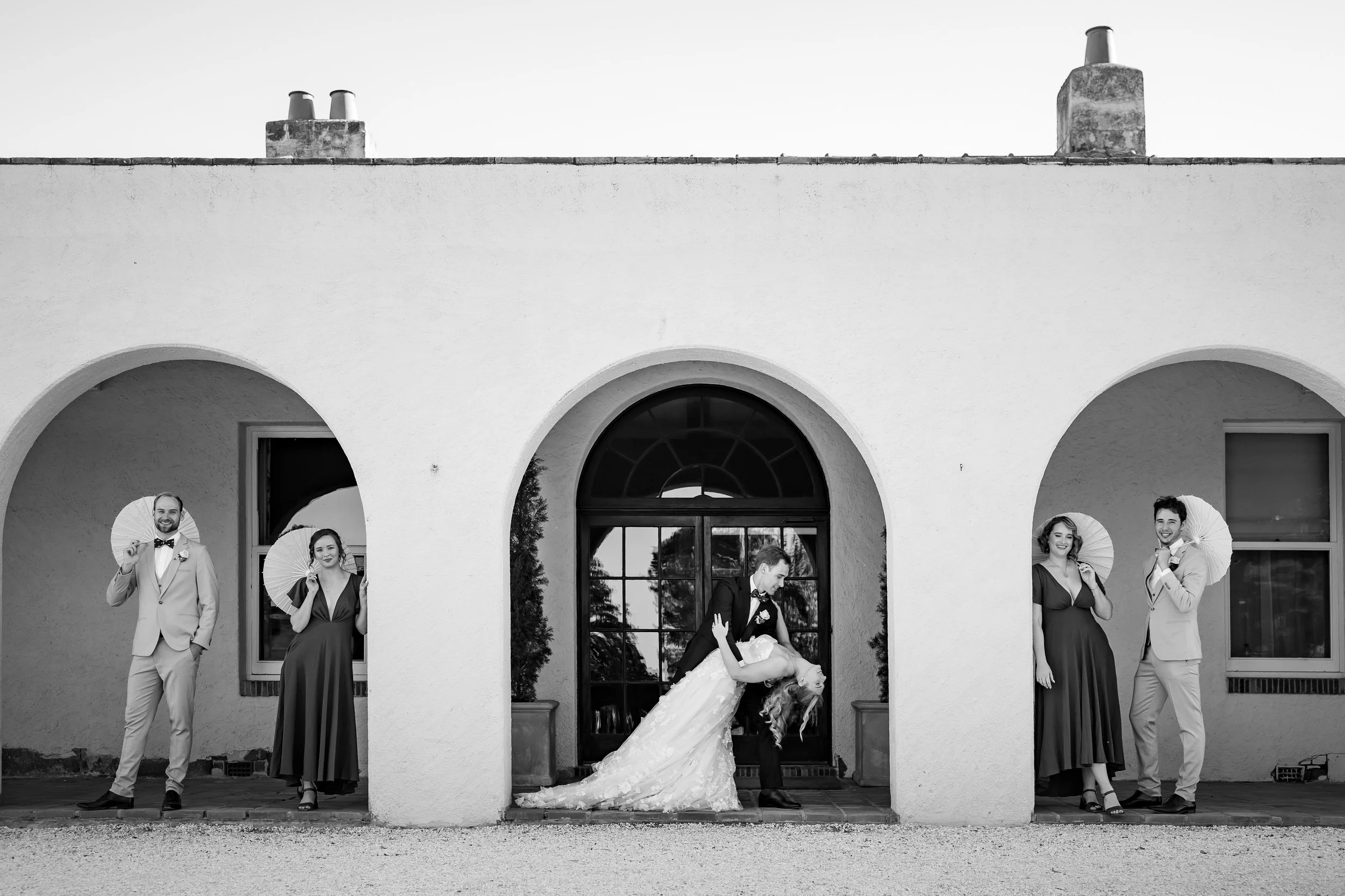 A group of six people dressed in formal attire, the women wearing evening gowns and the men in suits, standing in a line under an archway of a building. They are holding paper parasols and posing for a picture, with some smiling and others looking at each other. A bride and groom are dancing in front of the group, with the groom dipping the bride while she smiles and leans backward.