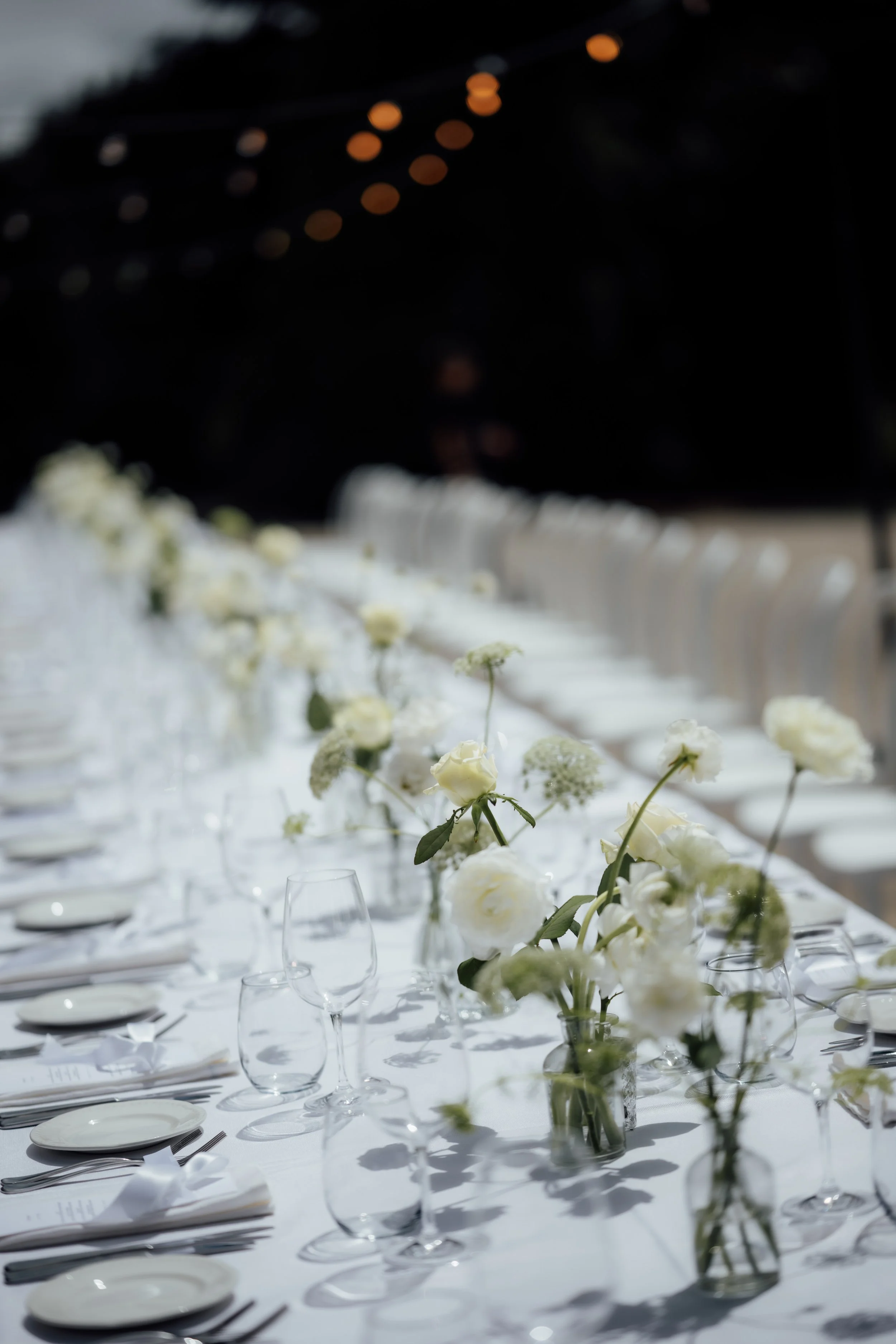 A long, elegant banquet table decorated with white flowers in glass vases. The table is set with white plates, silverware, and wine glasses under a dark ceiling with string lights.