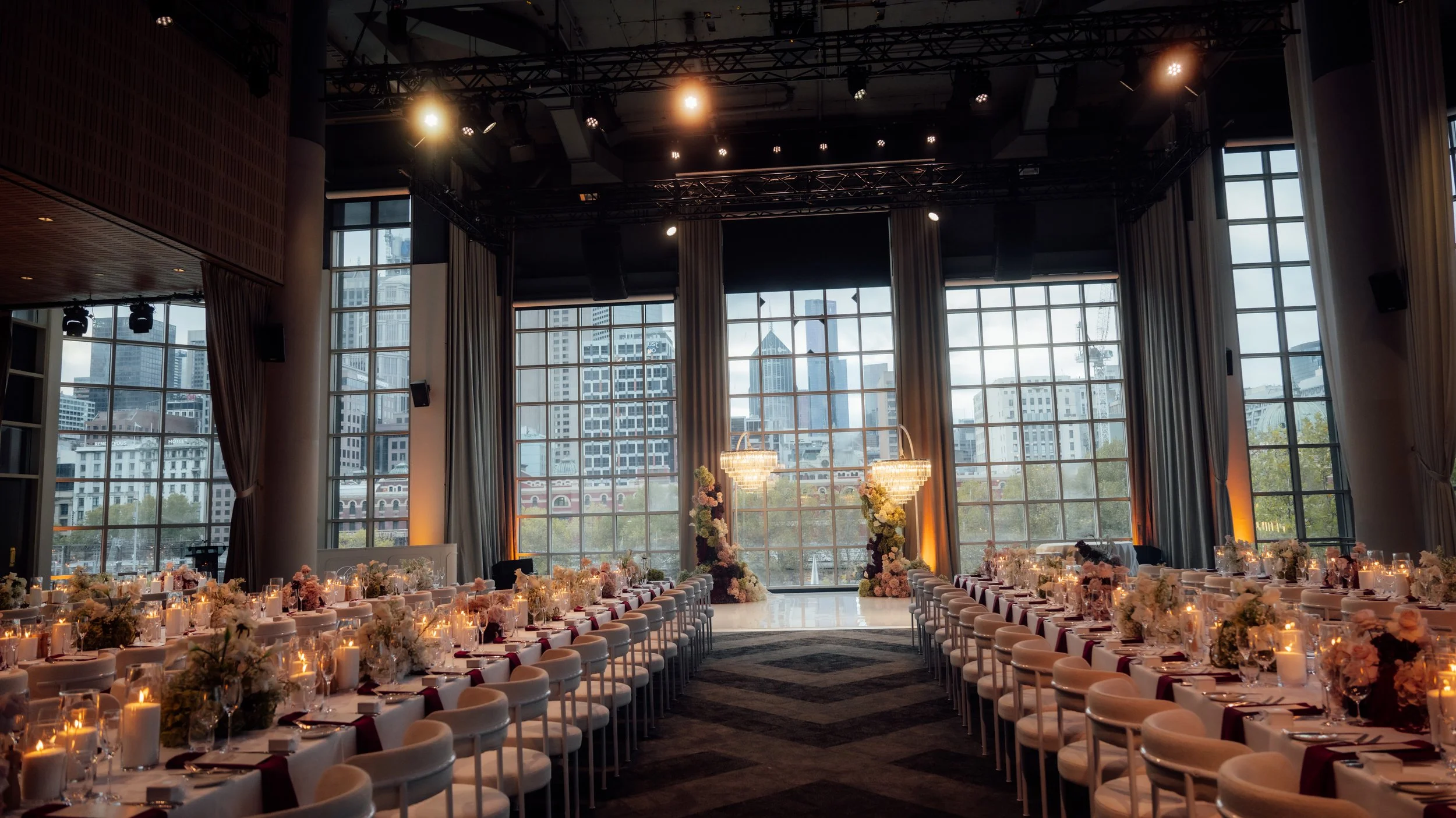 Elegant indoor event space with large windows, decorated for a wedding or banquet, featuring rows of tables with floral centerpieces and candles.