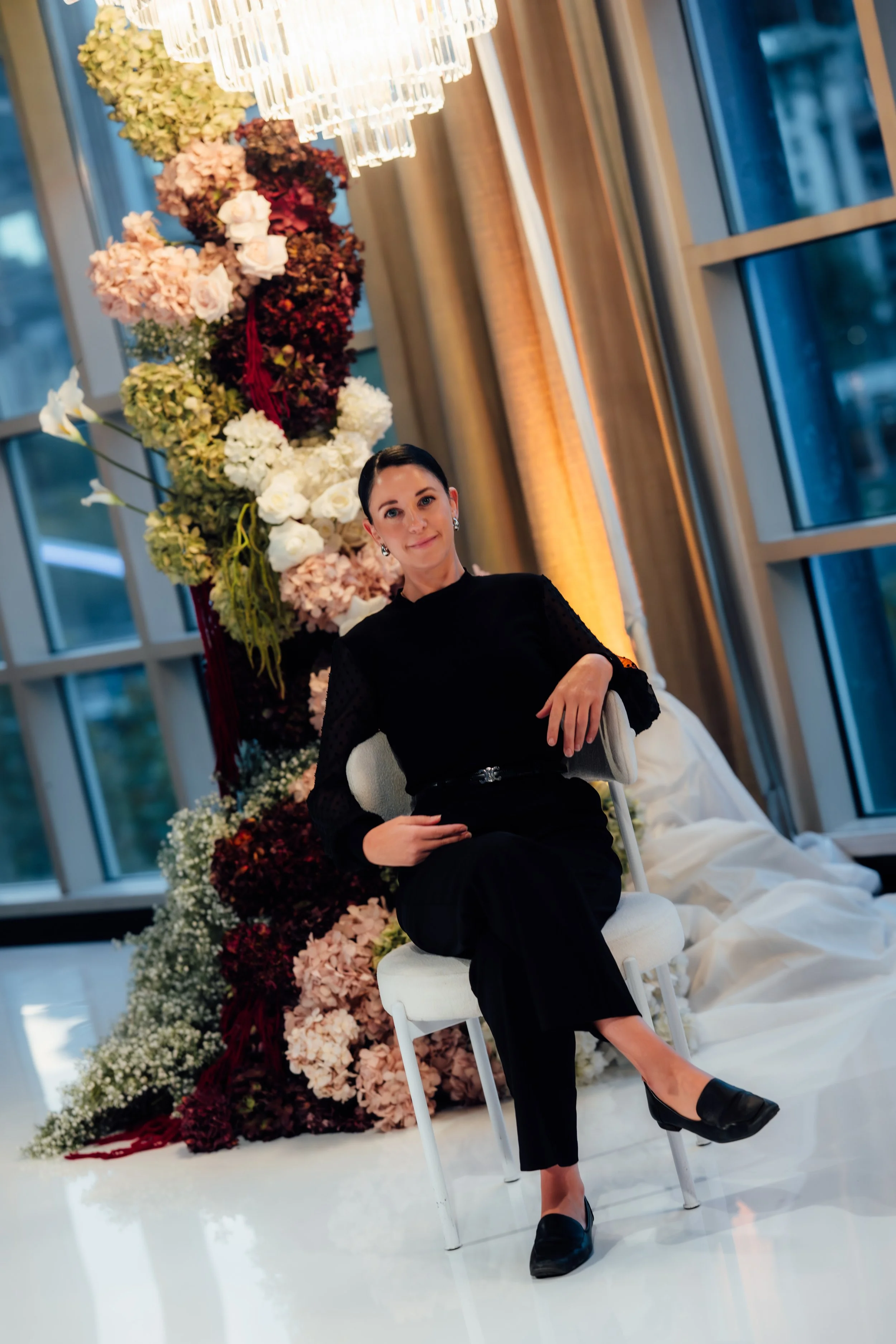A woman with short dark hair, wearing a black top and black pants, sitting on a white chair in front of a floral backdrop with an abundance of pink, white, and burgundy flowers. The setting appears to be indoors with large windows and warm lighting.
