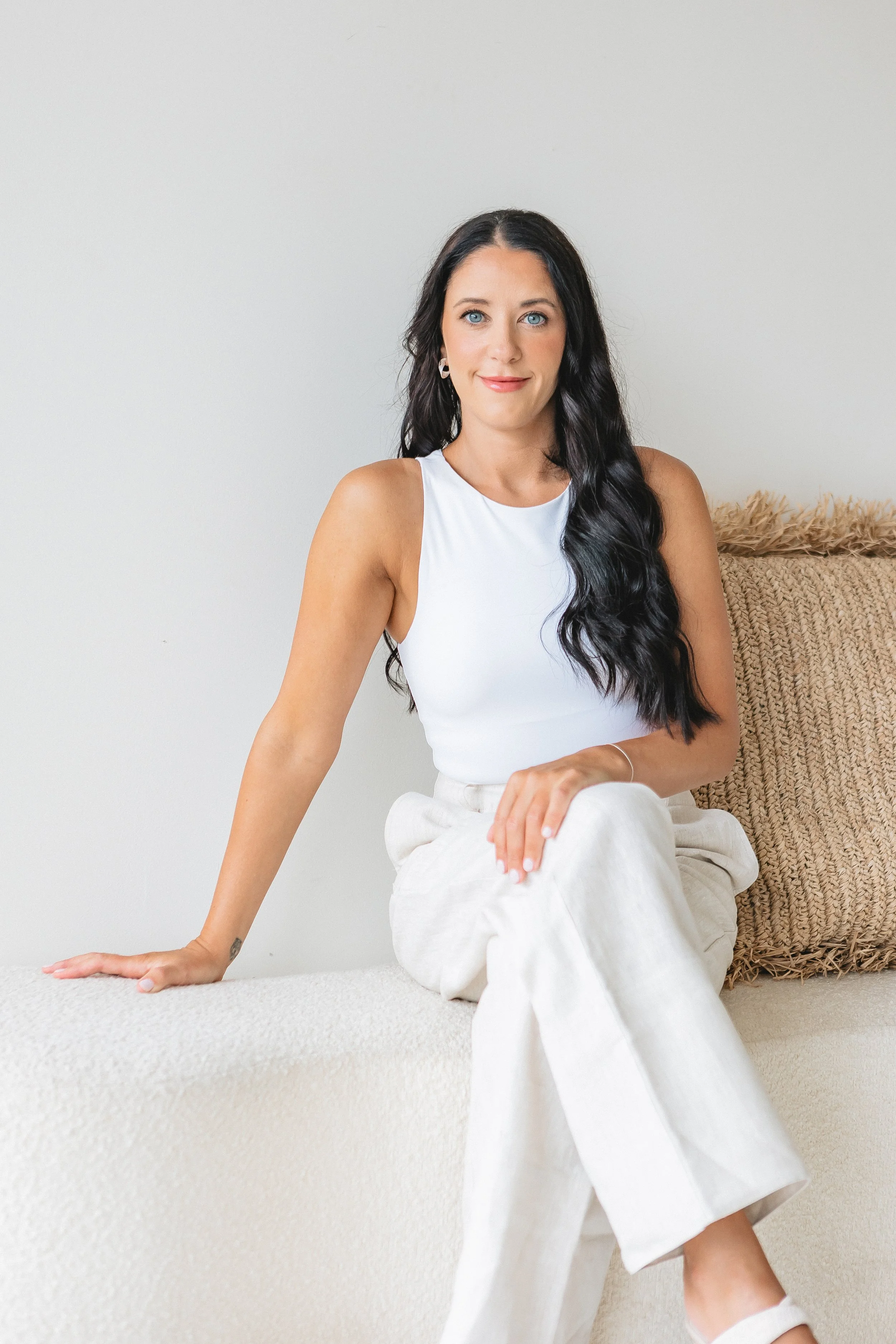 A woman with long black wavy hair, wearing a white sleeveless top and beige pants, sitting on a beige carpeted surface, with her right hand resting on the surface and her left hand on her knee, in a minimalist room with a plain light gray wall and a textured beige pillow behind her.