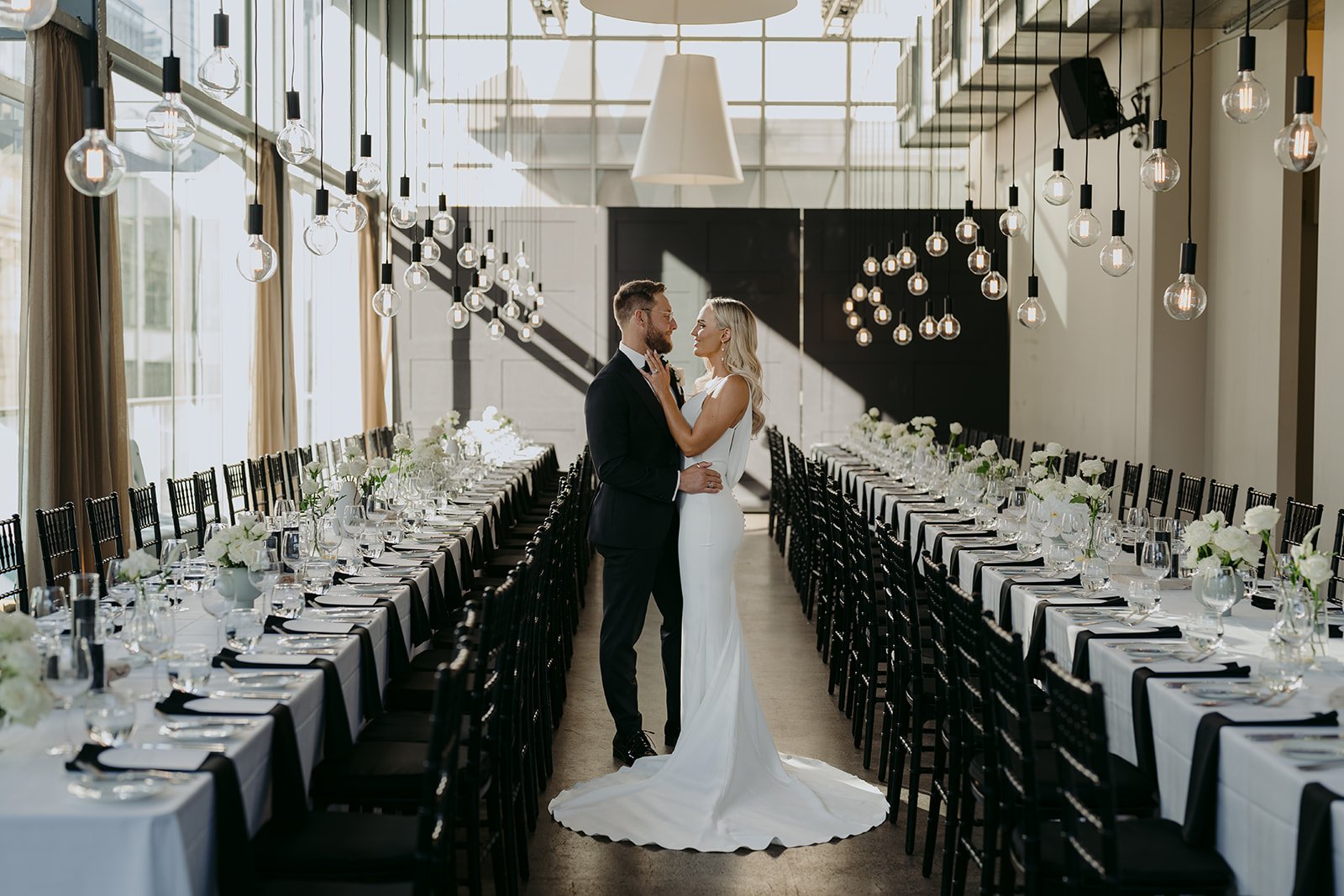 A bride and groom share a dance in a modern, well-lit wedding reception hall with long banquet tables decorated with white flowers, glassware, and black napkins. The hall has large windows and hanging light bulbs.