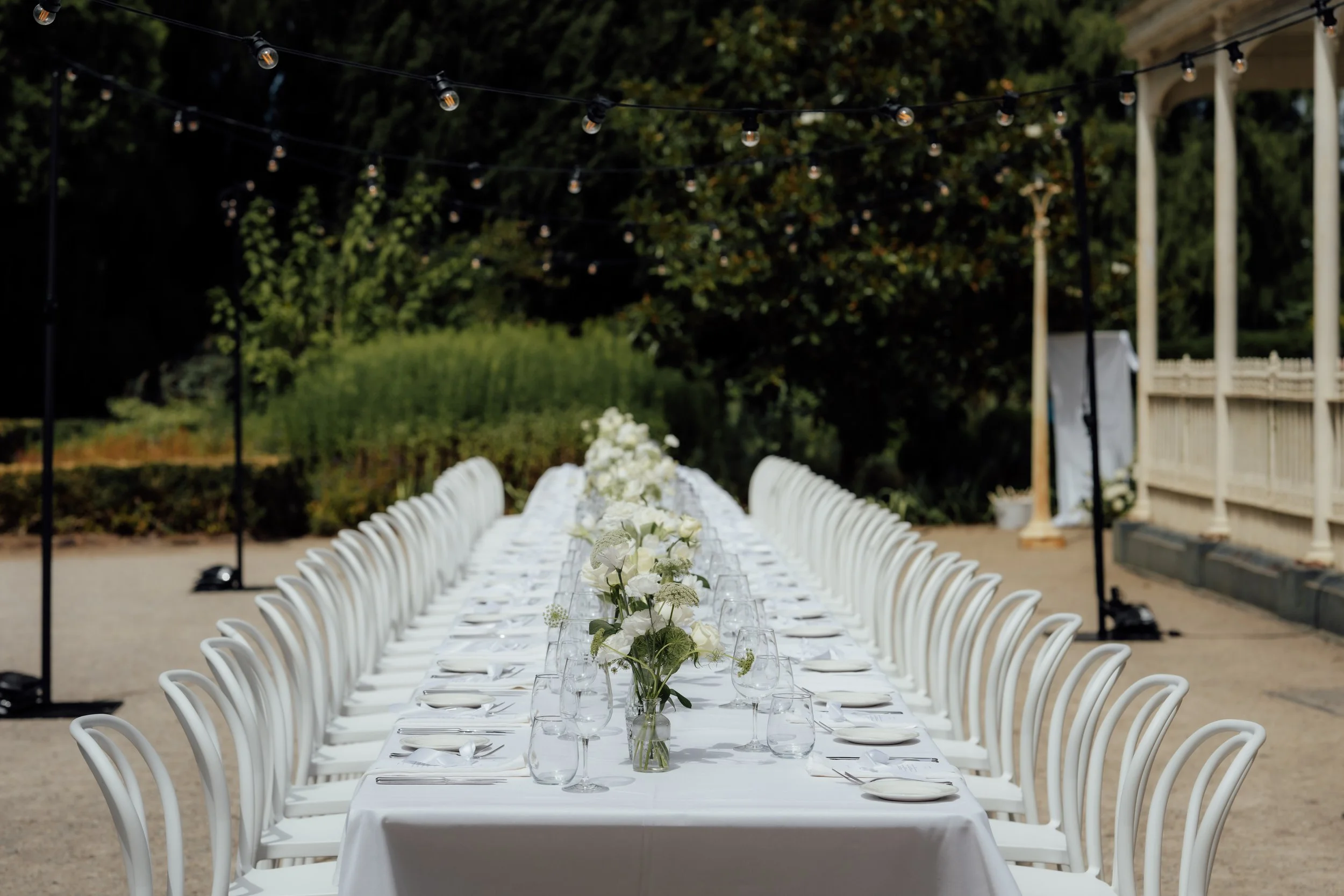 Outdoor wedding reception setup with a long white table, floral centerpiece, and string lights overhead, surrounded by chairs and greenery.