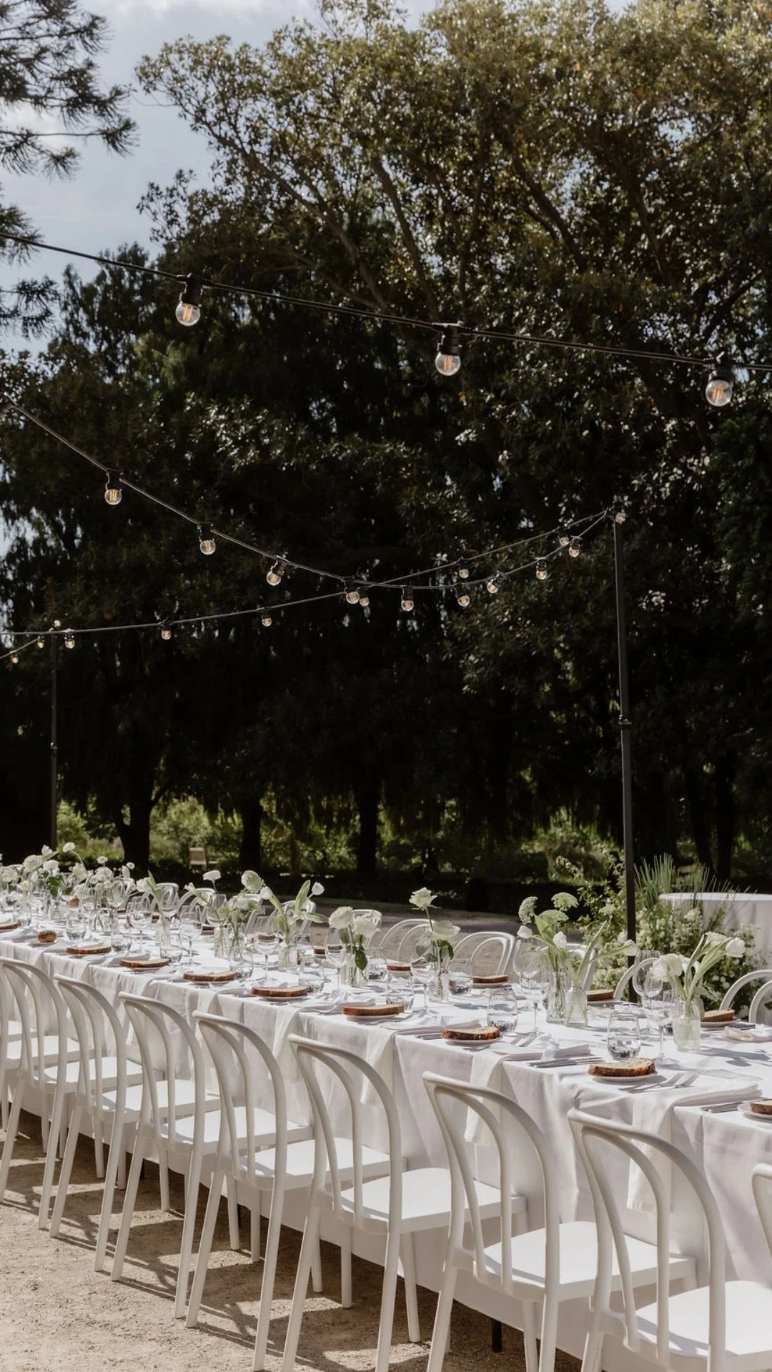 An outdoor dining setup with a long white table, white chairs, floral centerpieces, and string lights hanging above, surrounded by trees.
