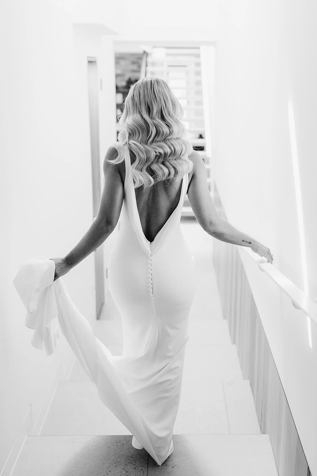 A woman in a backless wedding dress walking down a brightly lit hallway.