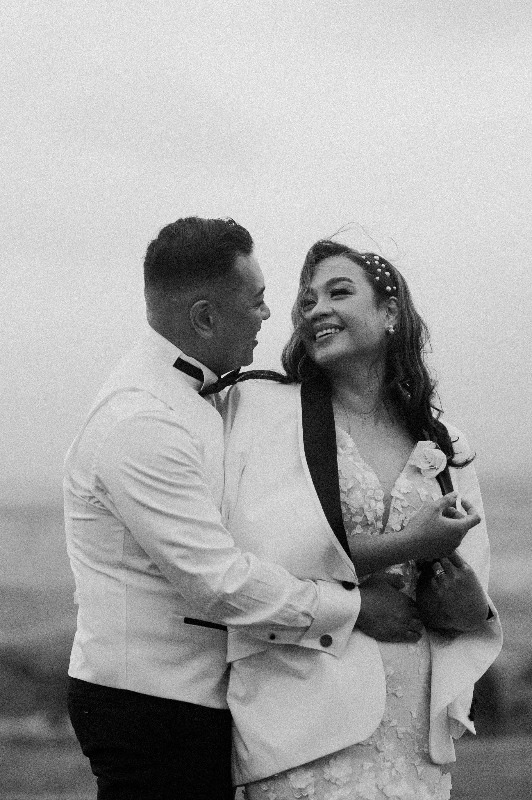 A black-and-white photograph of a couple dancing and smiling at each other, dressed in formal wedding attire.