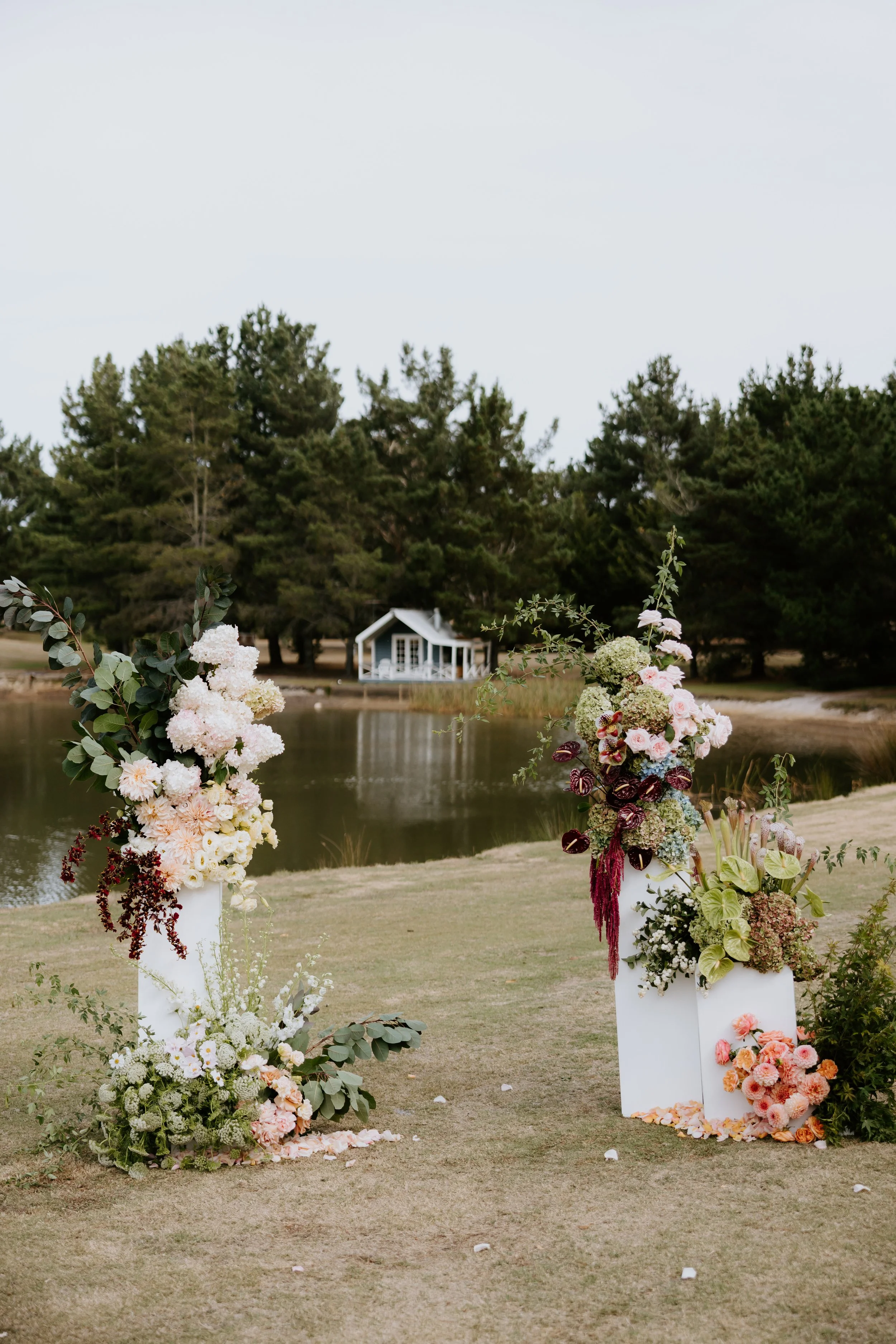 Outdoor wedding setup with floral arrangements on white pedestals near a small lake, with trees and a cabin in the background.