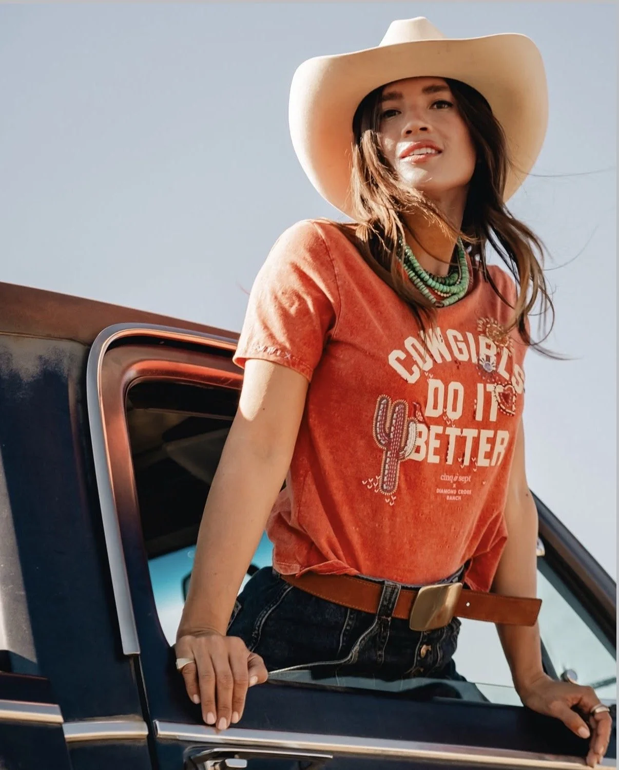 A woman wearing a cowboy hat, orange t-shirt, layered necklaces, and jeans, leaning out of a pickup truck against a clear blue sky.