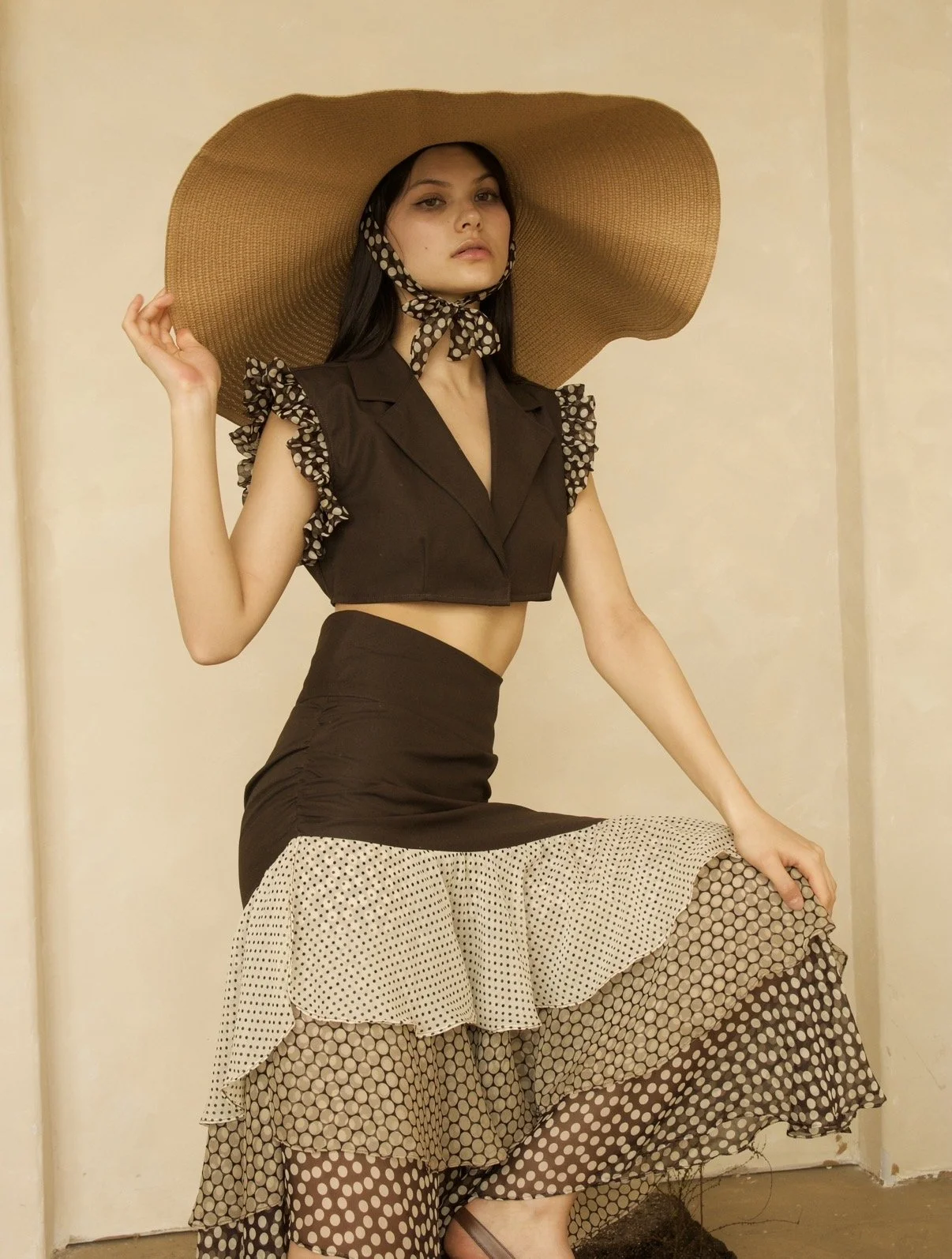 A woman wearing a large brown sun hat, a black sleeveless crop top with ruffled polka-dot details on the shoulders, and a layered polka-dot skirt, standing against a plain wall.