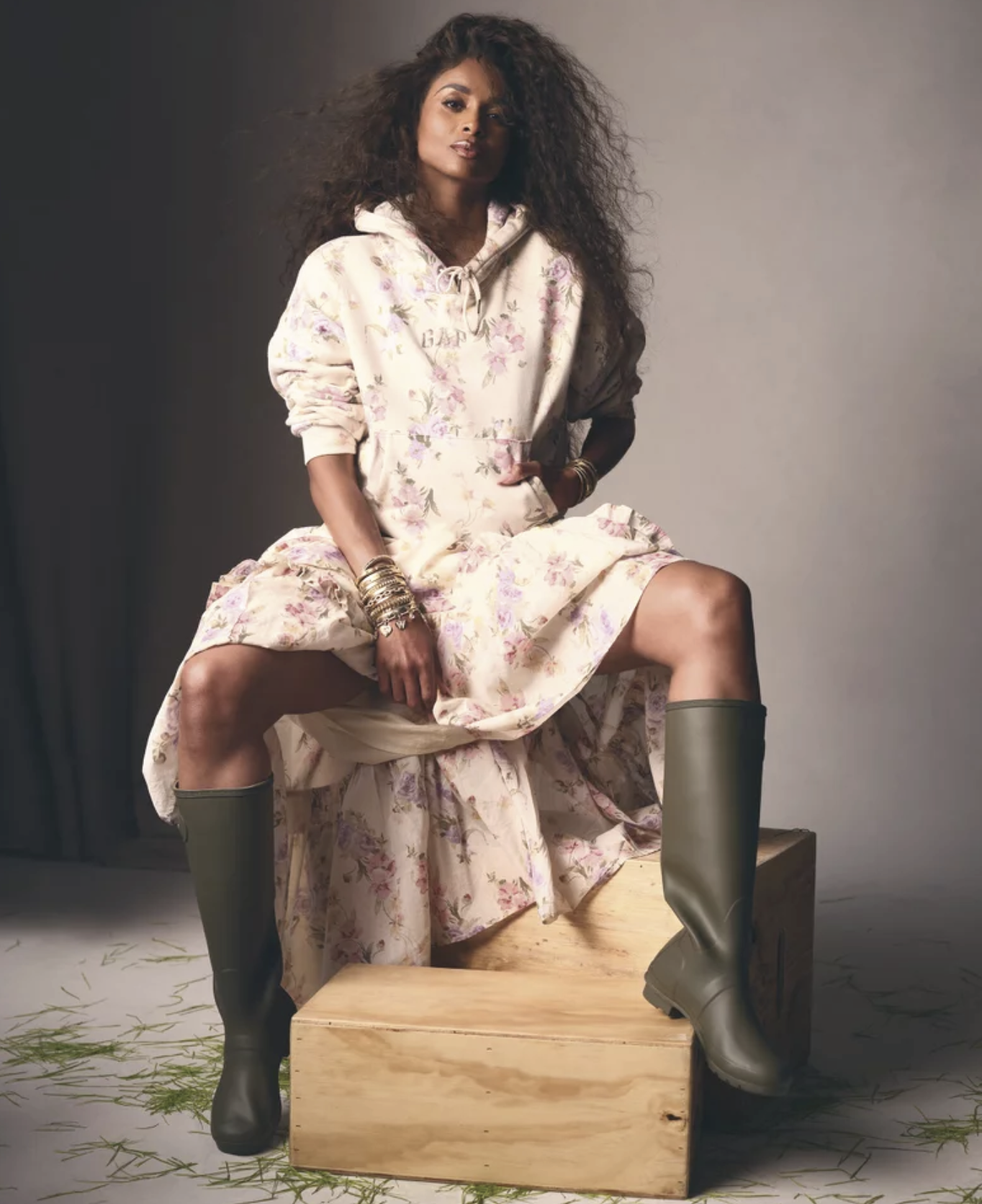 A woman with curly hair wearing a floral hoodie and boots, sitting on a wooden box with her legs spread apart, with some grass scattered on the floor around her.