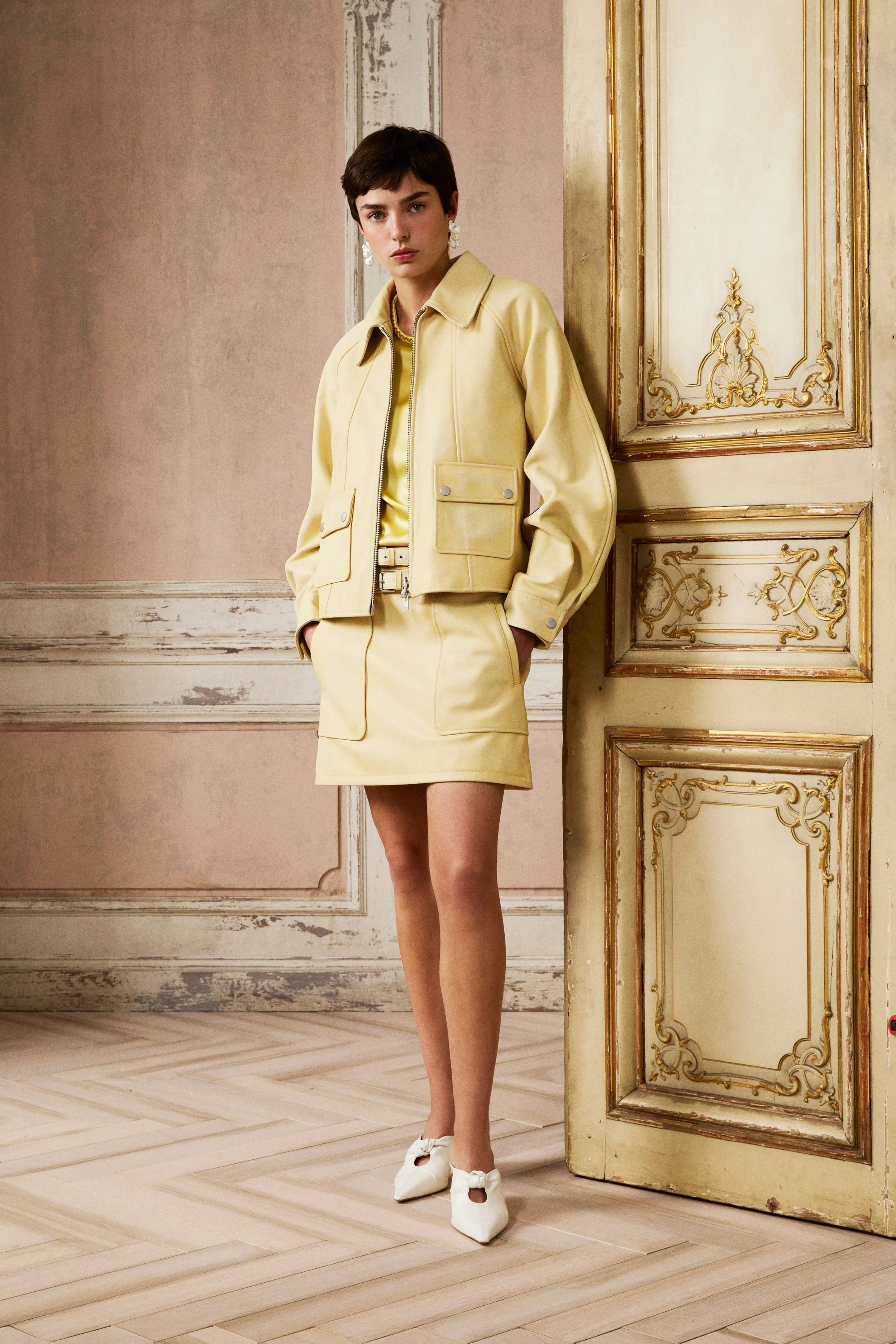 A woman with short dark hair wearing a yellow jacket and skirt, white shoes, standing in a vintage ornate room with decorated gold and cream-colored walls and parquet flooring.