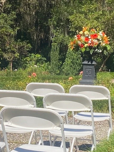 An outdoor setting with white folding chairs arranged in rows and a bouquet of colorful flowers on a pedestal in the background.