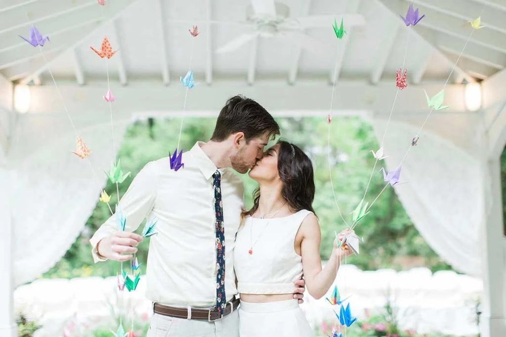 A couple kissing under hanging colorful origami cranes in an outdoor pavilion.