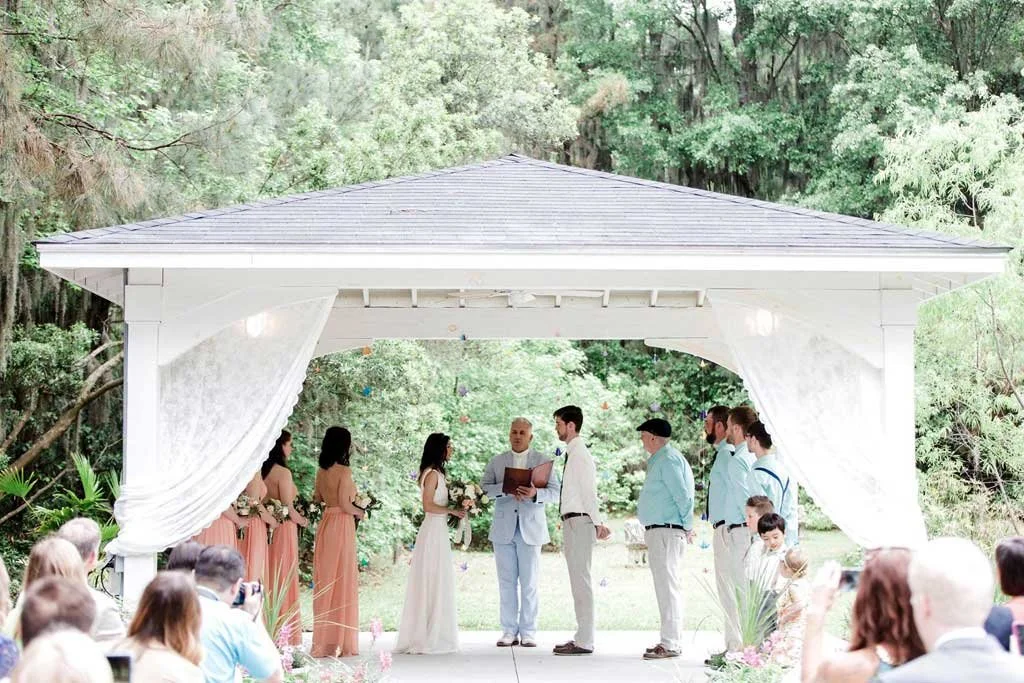 A wedding ceremony taking place outdoors under a white pavilion with curtains, surrounded by trees. The bride and groom are standing with their wedding party, and guests are seated watching.
