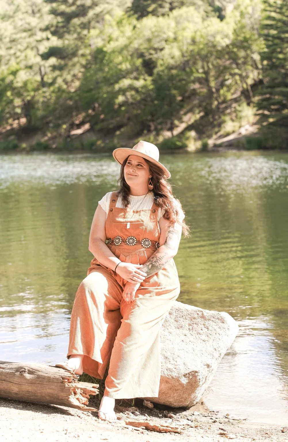 Claire Ramirez-Yocum, therapist at Rooted Pines Healing, seated on a boulder at the edge of a mountain lake.